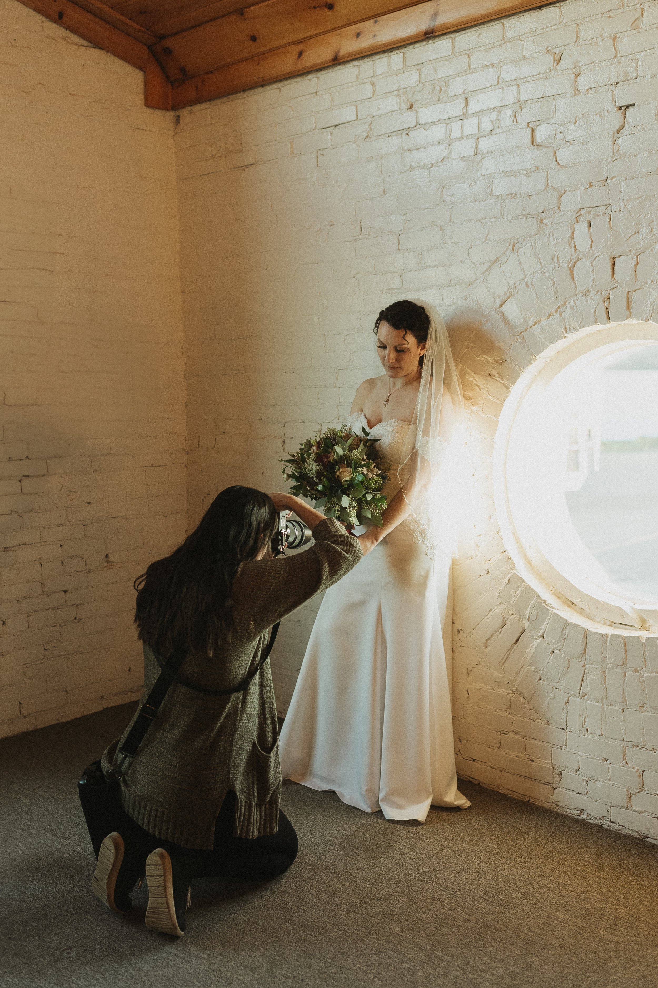 A bride in a white wedding dress with a veil holds a bouquet of flowers, while a photographer kneels to take her picture inside a room with white brick walls and a round window.