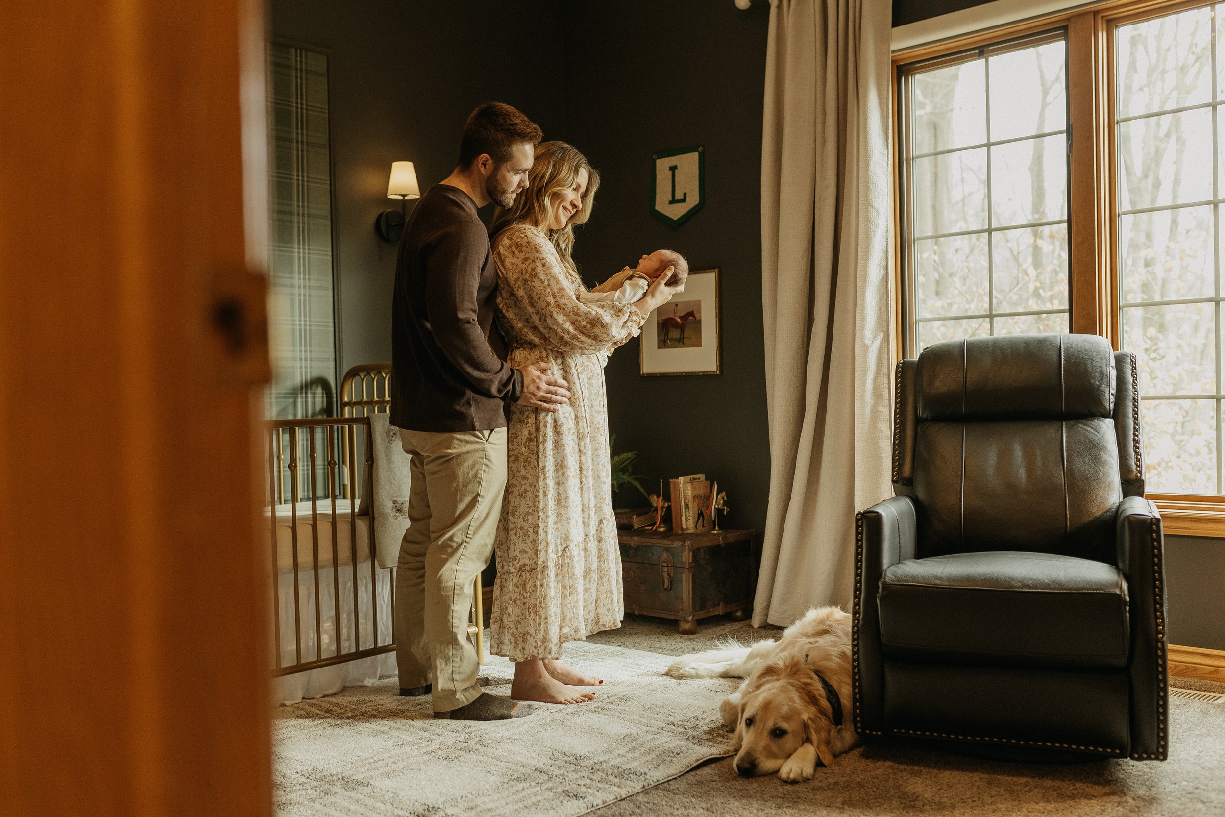 Couple standing in a cozy nursery, holding a baby. A dog is lying on the floor near a leather recliner. The room has a crib, dark walls, and a large window with curtains.