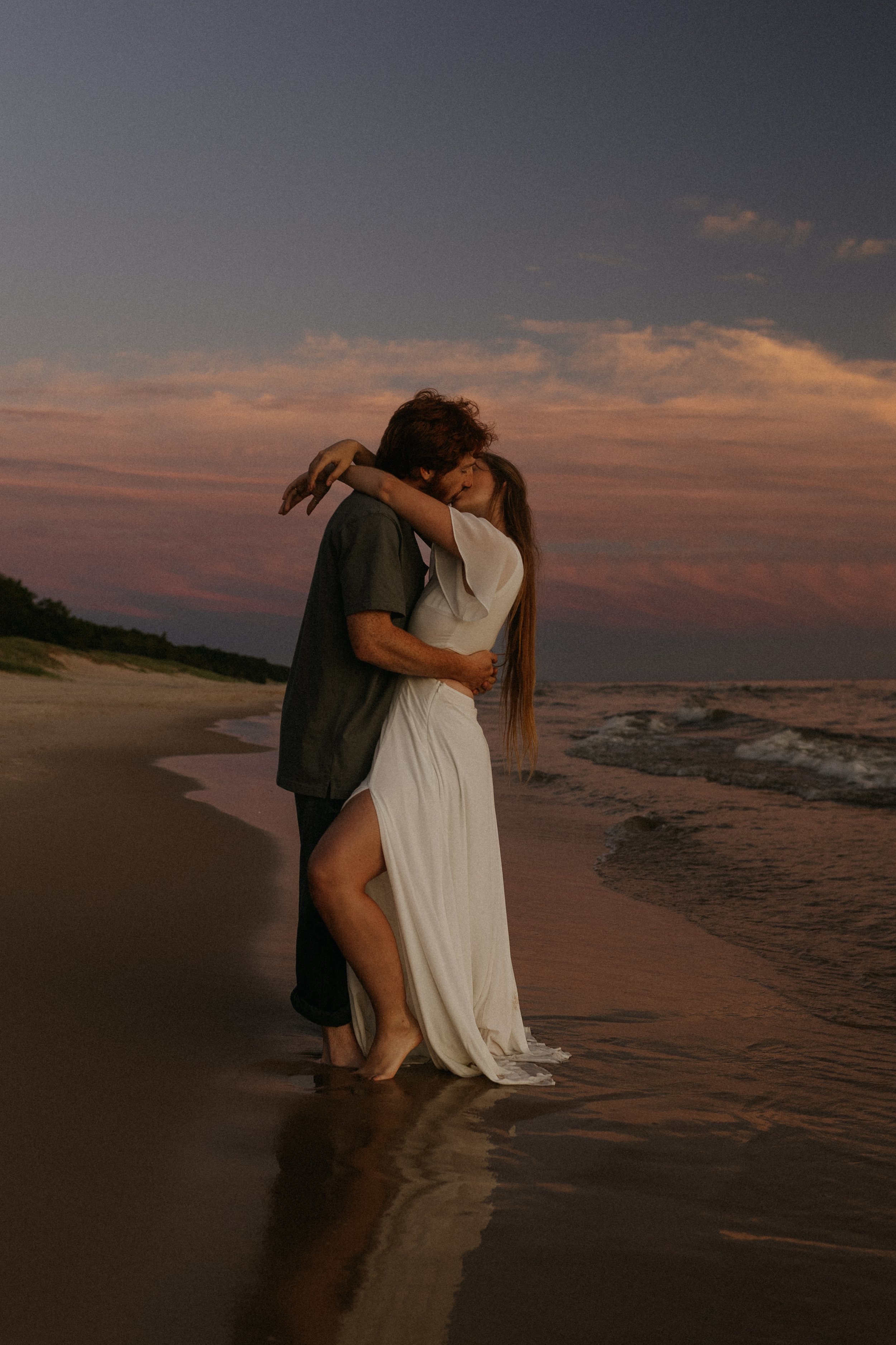 A couple kissing on a beach at sunset, with the woman in a white dress and the man in dark clothing, standing in shallow water.
