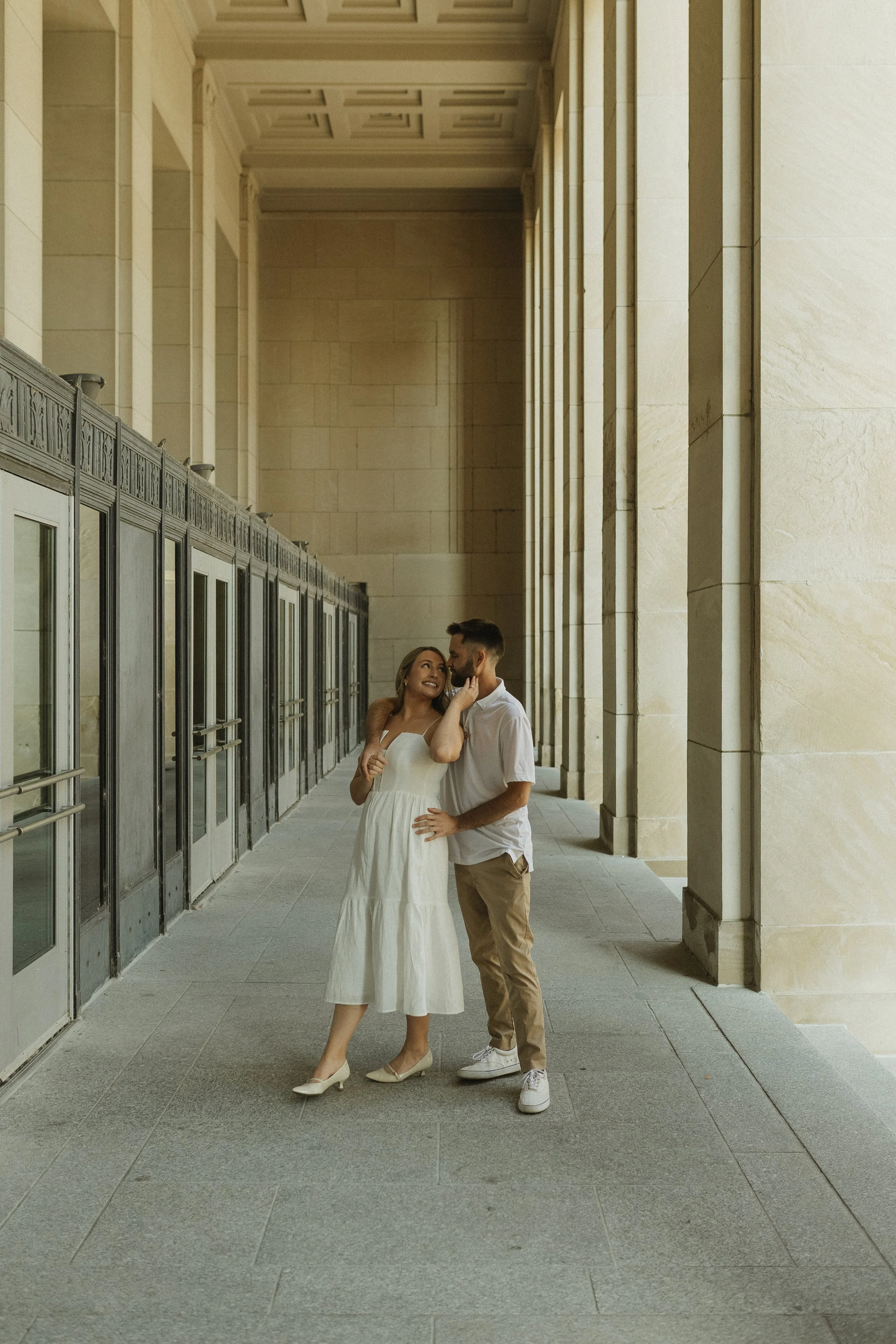 A couple sharing an intimate moment in an architectural corridor with tall columns and large glass-door entrances, both dressed in light-colored clothing.