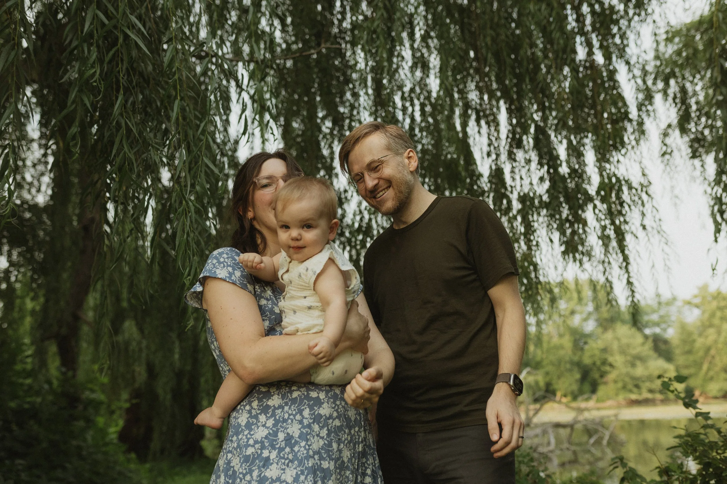 A happy family of three outdoors under a tree, with a woman holding a toddler and a man standing beside them, smiling.