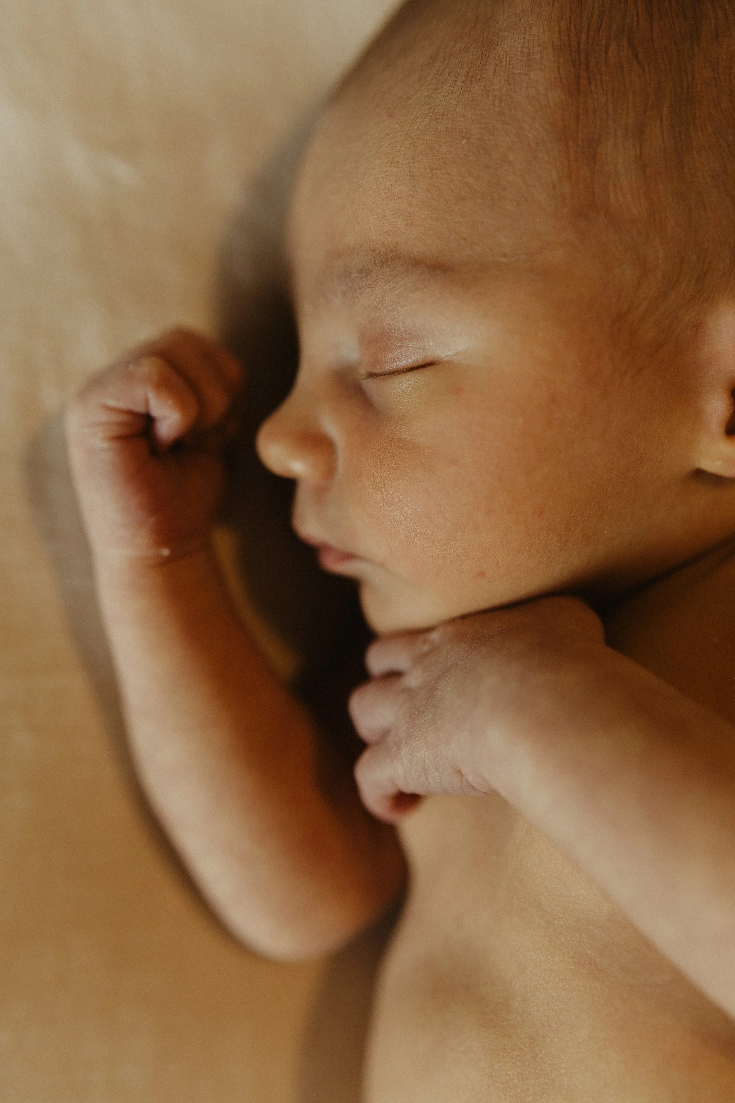 Close-up of a sleeping baby with eyes closed, hand near face, resting on a soft surface.