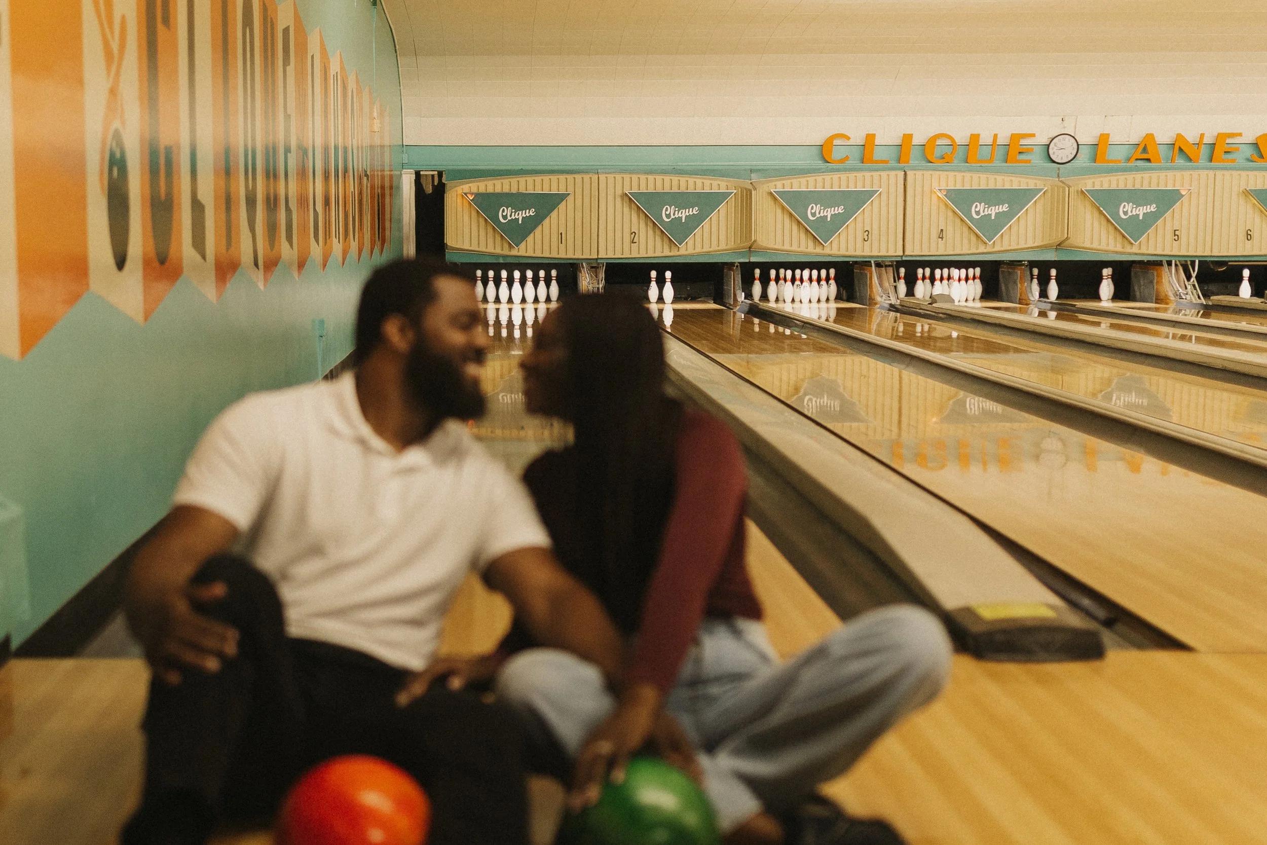 A couple sitting on the floor of a bowling alley, holding bowling balls and preparing to bowl.