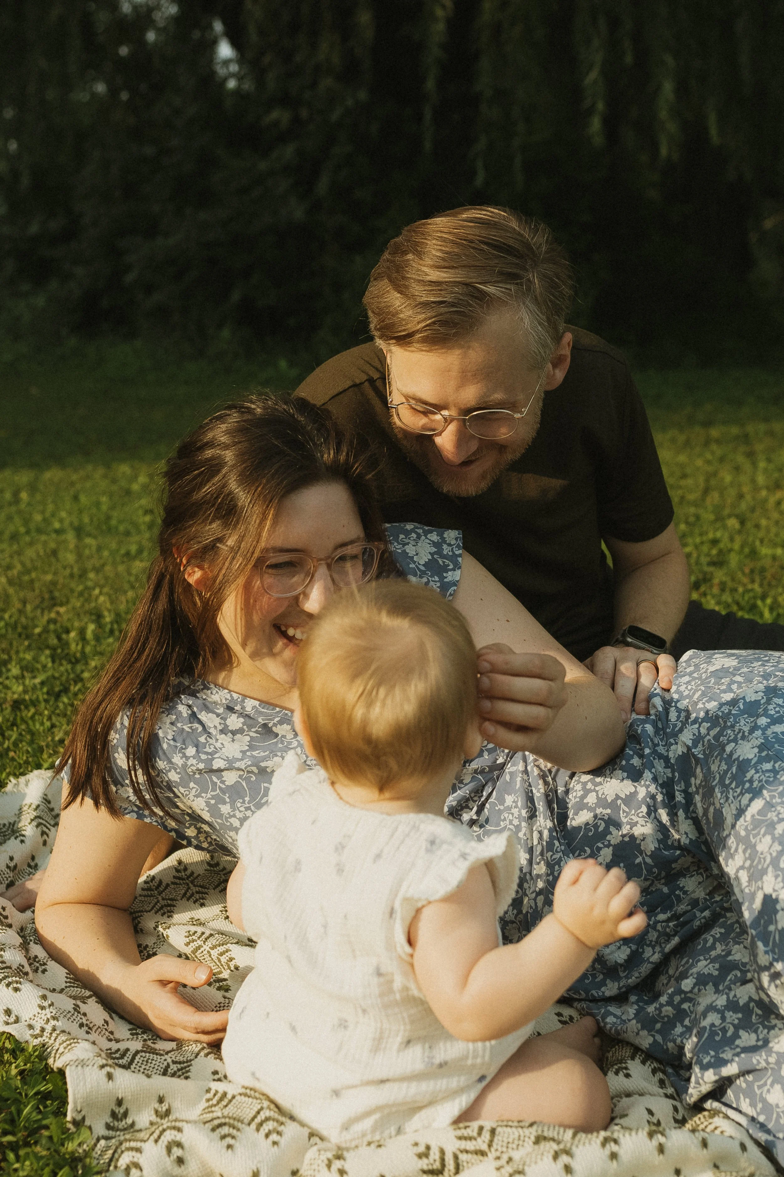 A family of three, including a woman, a man, and a baby, sitting on a blanket outdoors, enjoying a sunny day. They are smiling and interacting, with the woman and man focusing on the baby.