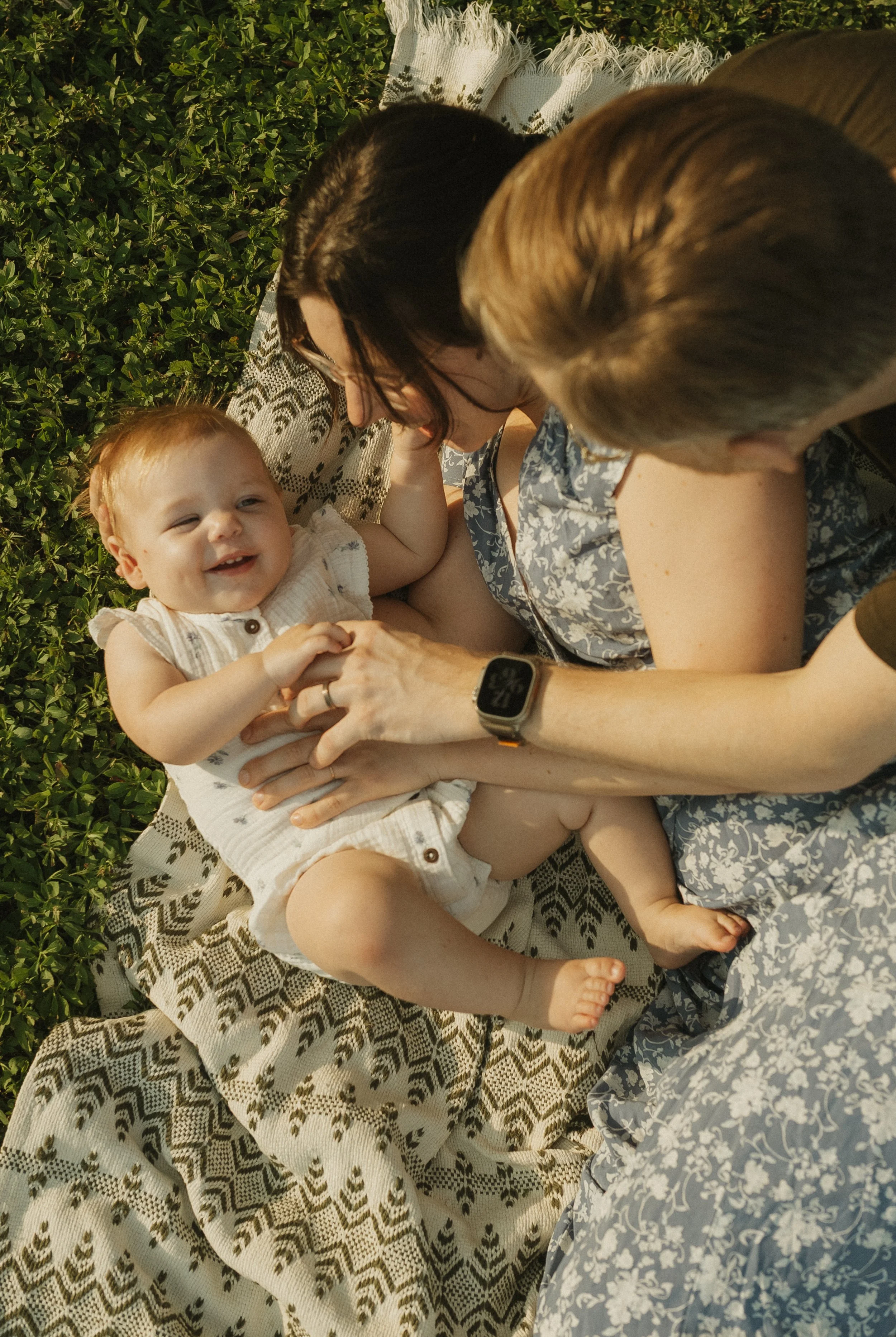 A woman, a man, and a baby lying on a patterned blanket outdoors surrounded by greenery. The baby is smiling and looking at the woman, who is holding the baby's hands, while the man is leaning towards them.
