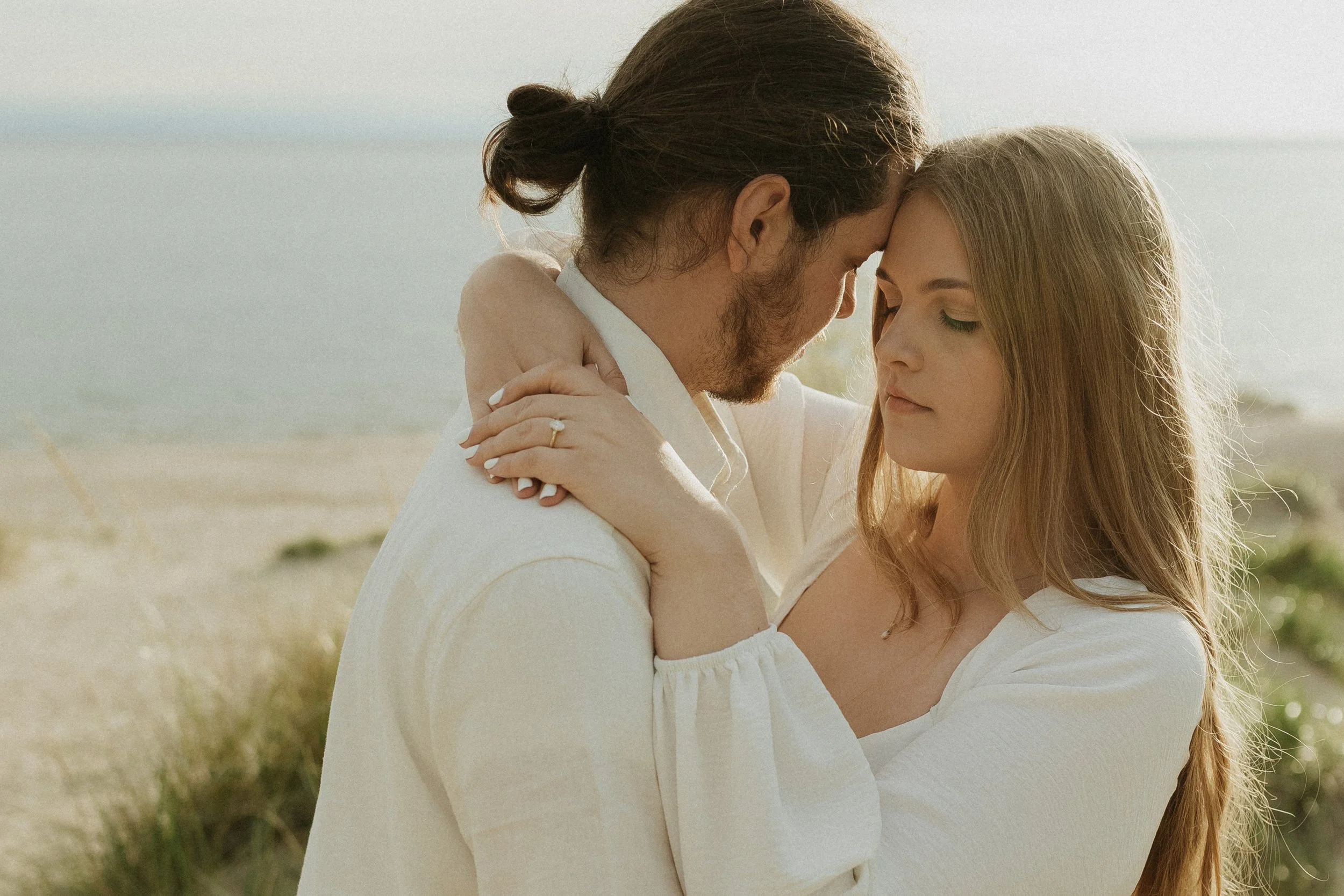 A couple embraces on a beach at sunset, with their foreheads touching and eyes closed.