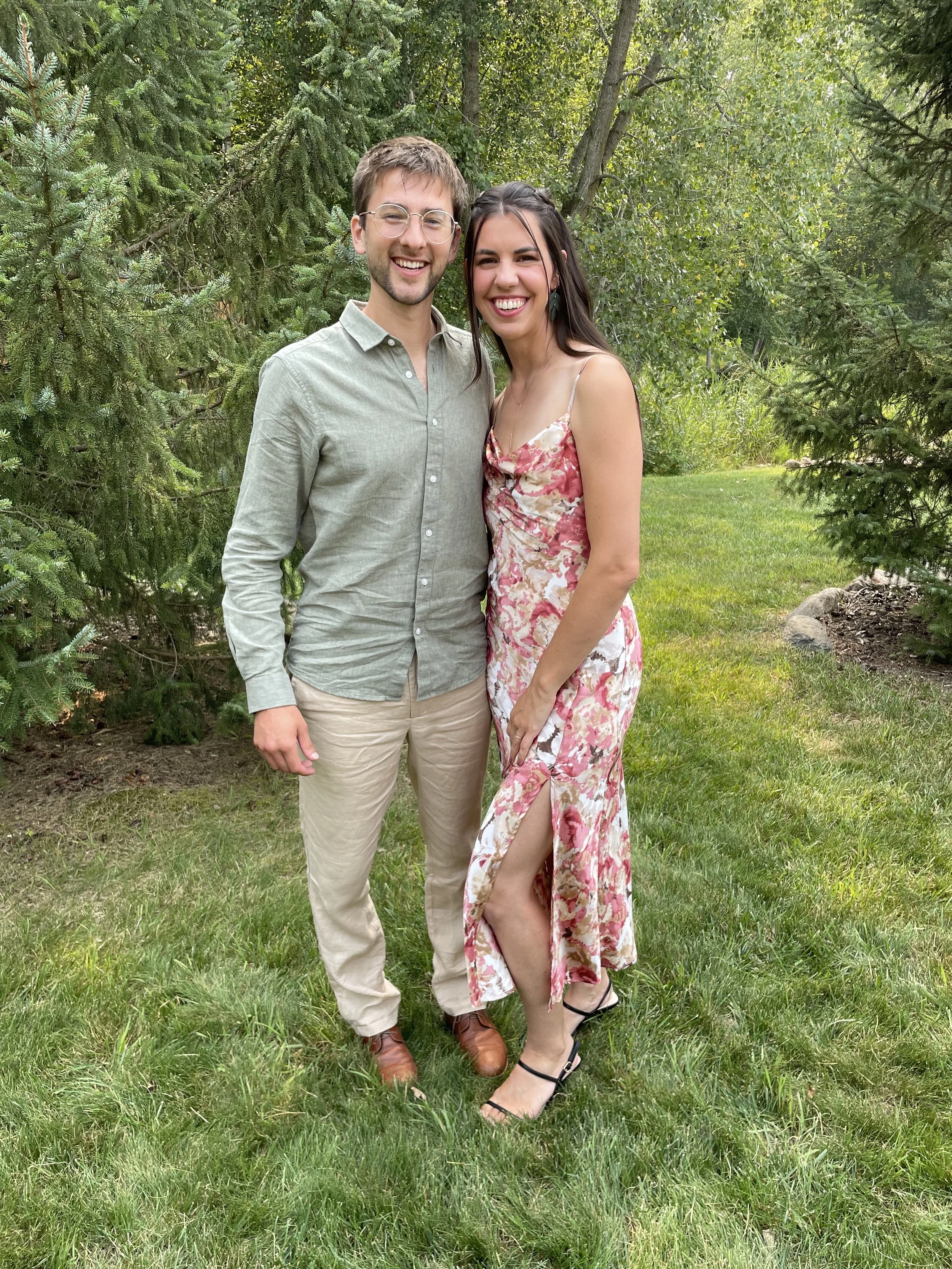 A smiling young man and woman standing side by side outdoors in a garden surrounded by green trees and grass. The man wears glasses, a light green button-up shirt, beige pants, and brown shoes. The woman wears a pink and white floral dress with a sli