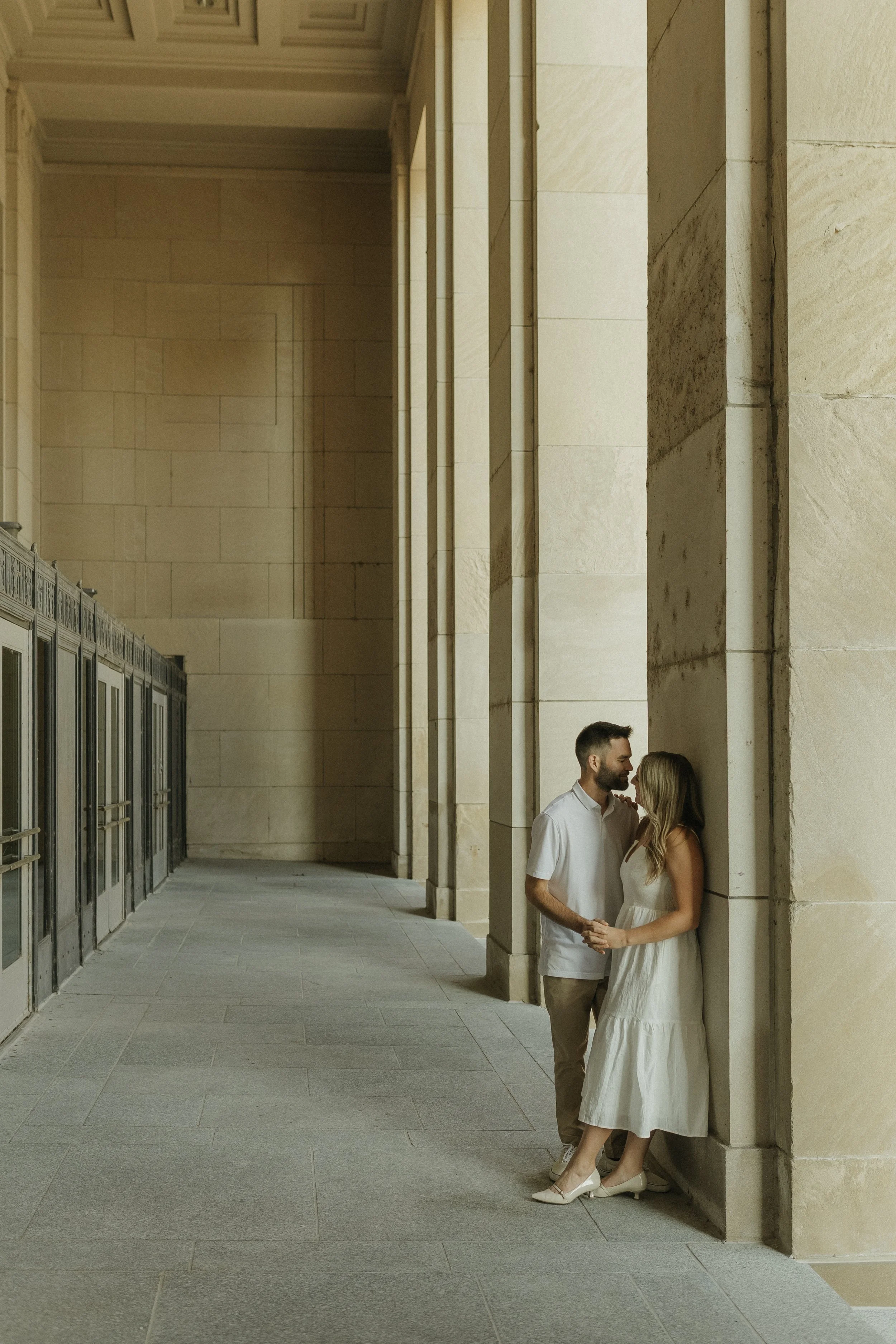 A couple standing close, holding hands, and leaning against a large stone column inside a building with tall walls and a high ceiling.