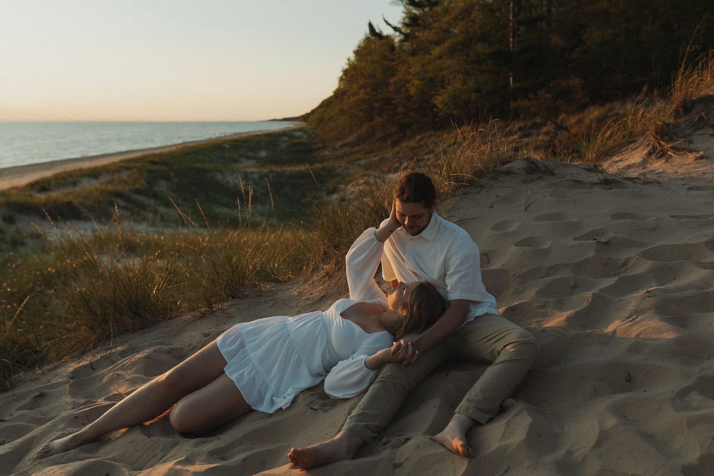 A couple sharing an intimate moment on a sandy beach during sunset, with the woman lying down and the man sitting up, holding her hand and looking at her.