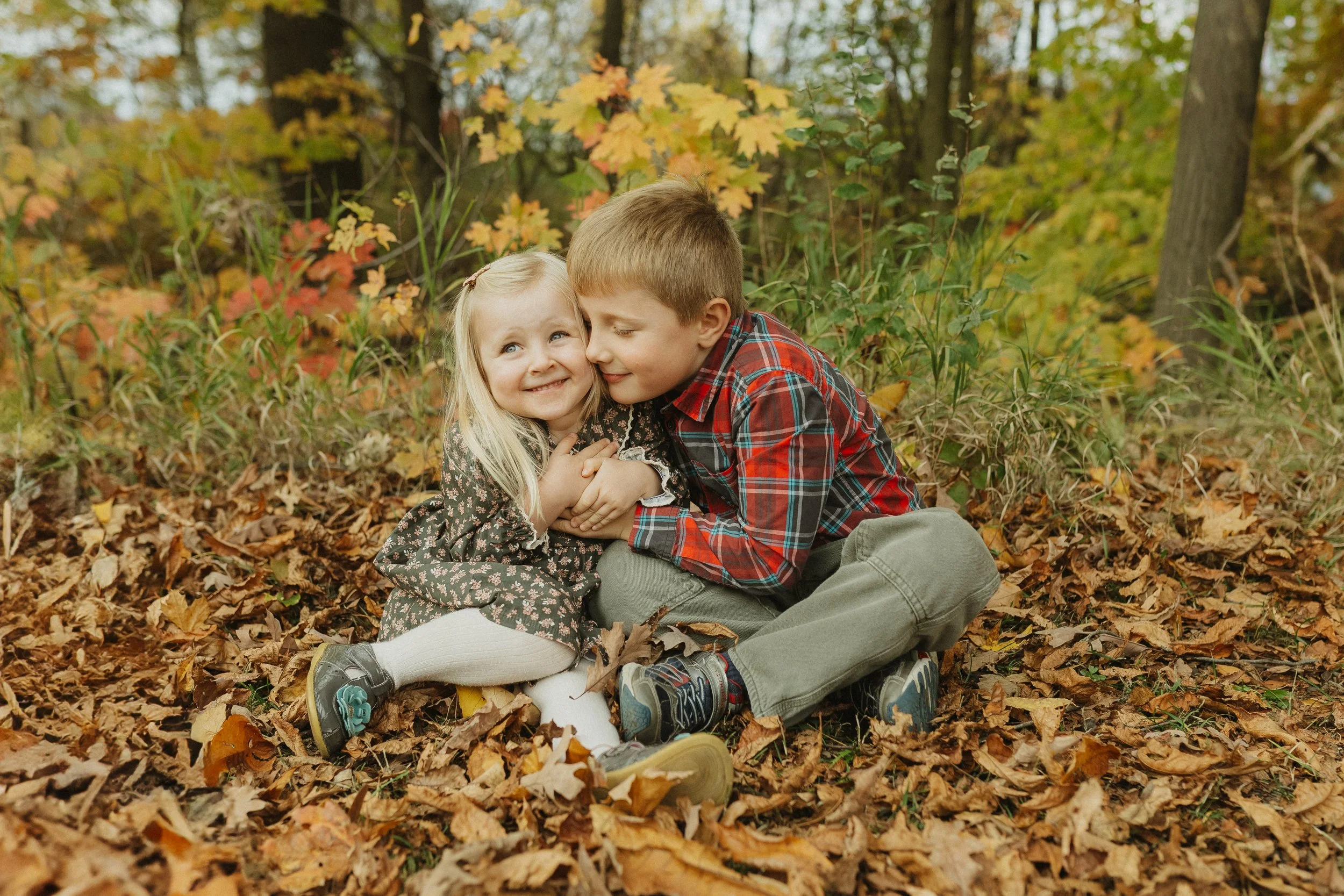 A young boy and girl sitting on fallen autumn leaves in a wooded area, hugging and smiling at each other.