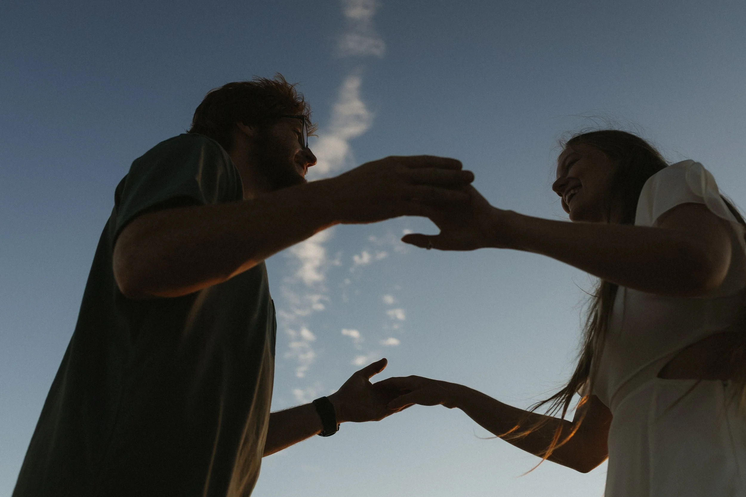 A silhouette of a man and woman facing each other, touching hands with the sky and clouds in the background during sunset.