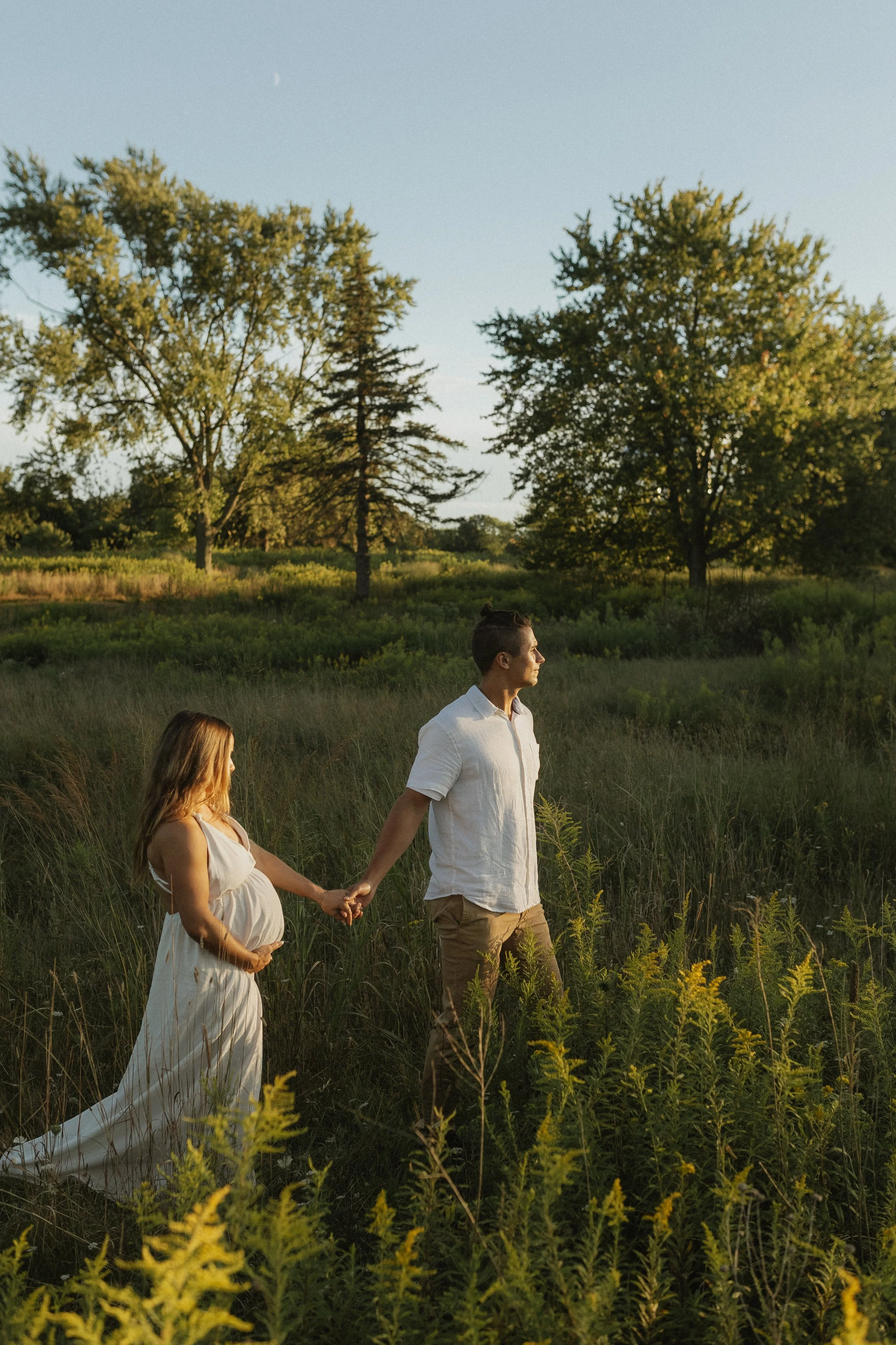 A pregnant woman and a man holding hands in a grassy field with trees, during sunset.