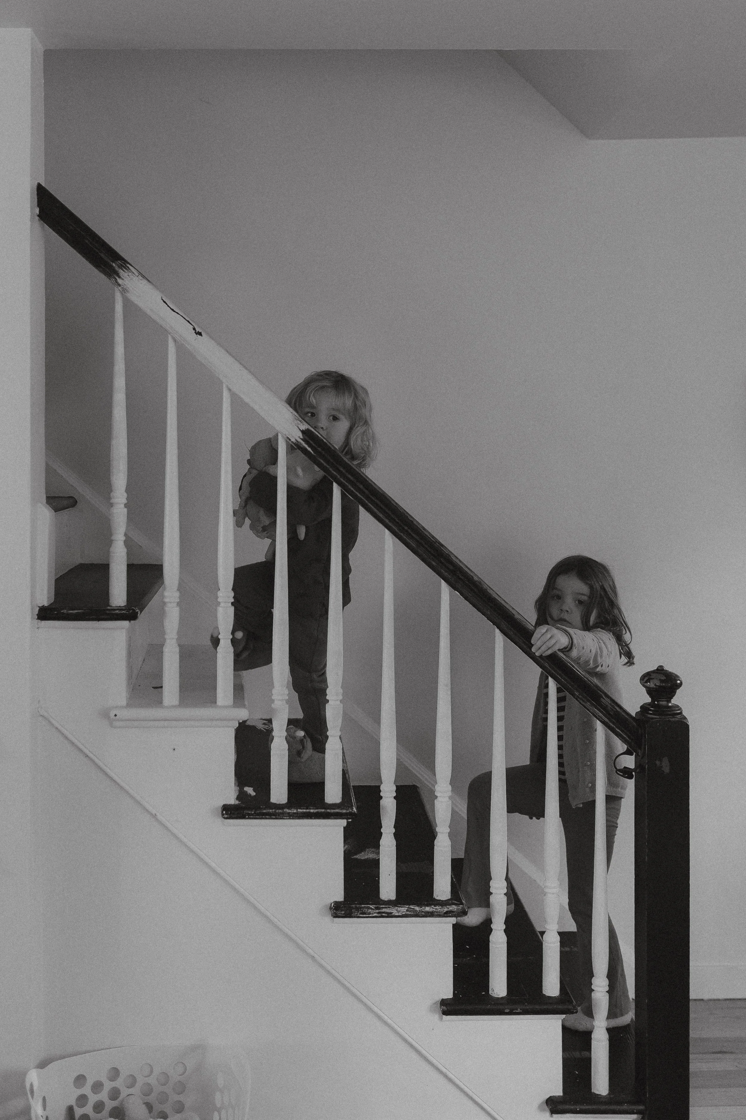 Two children standing on a staircase, holding onto the railing, one looking directly at the camera, in a house with plain walls.