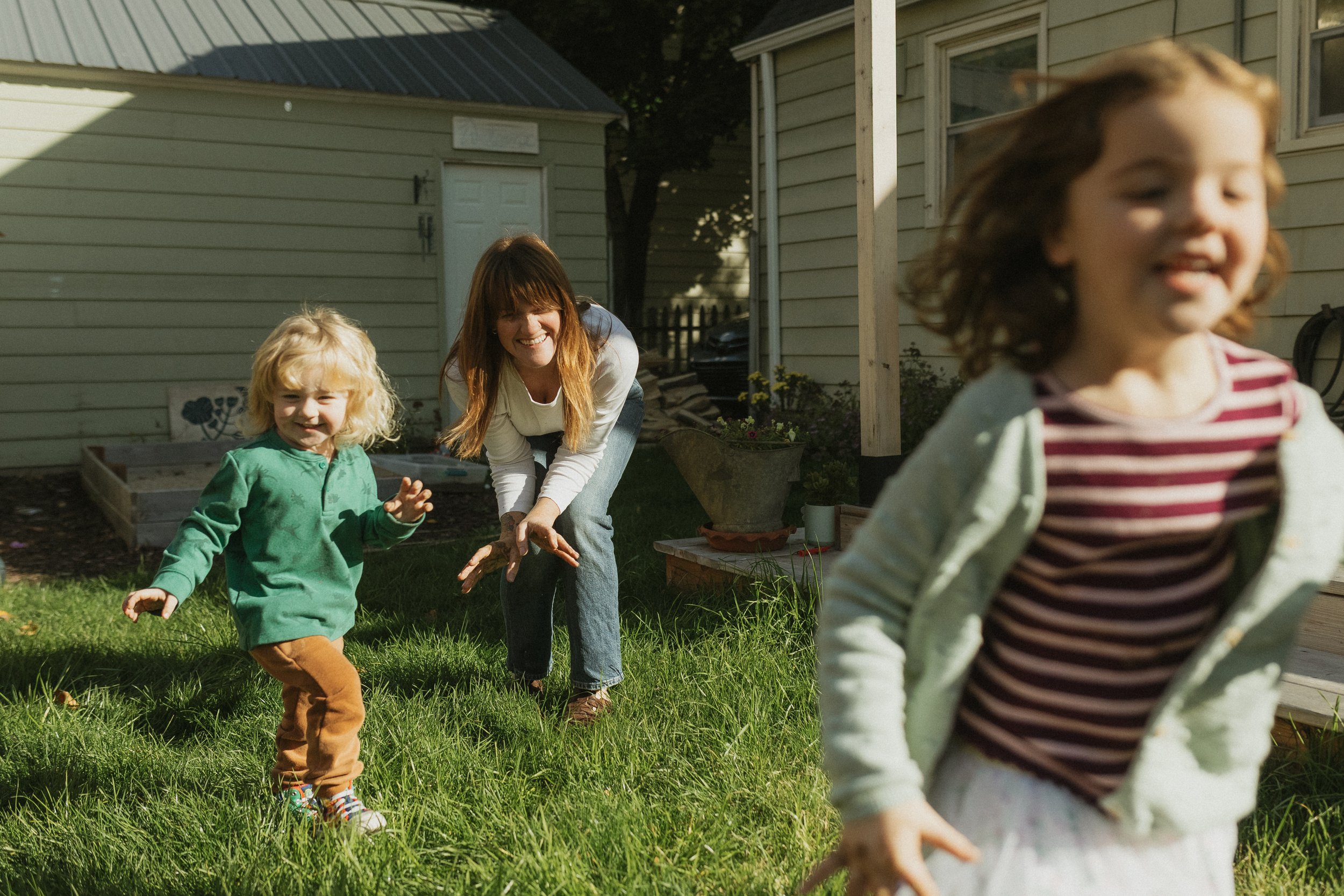 A woman and two young girls playing tag outdoors in a backyard on a sunny day.