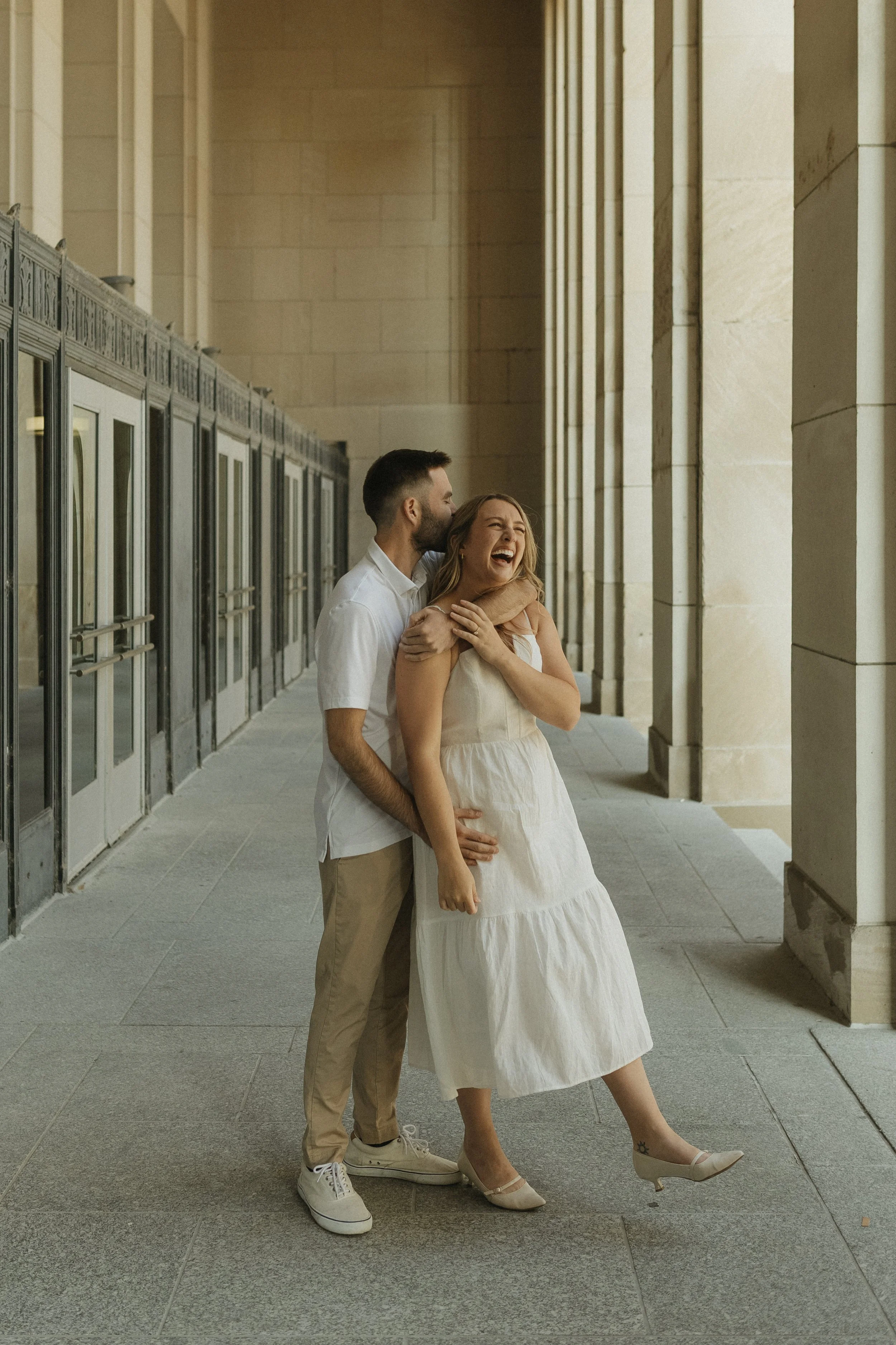 A man and woman laughing and embracing each other on a covered outdoor walkway with large stone columns and glass doors.