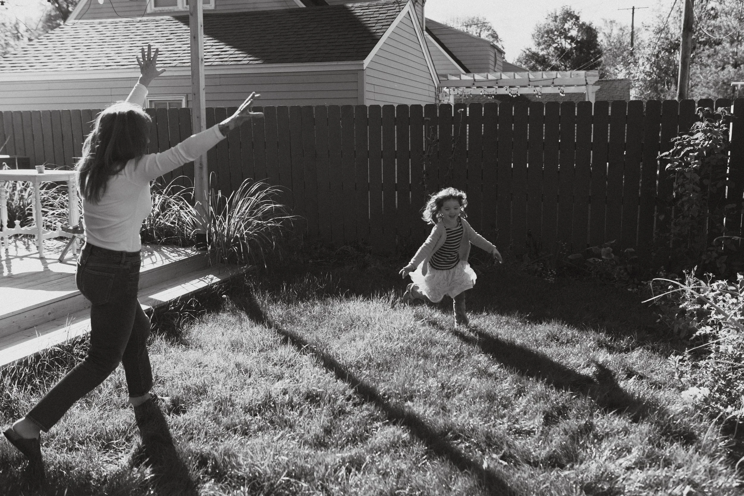 A woman and a young girl playing in a backyard with sunlight casting long shadows. The woman appears to be chasing the girl, who is running happily. The scene is in grayscale.