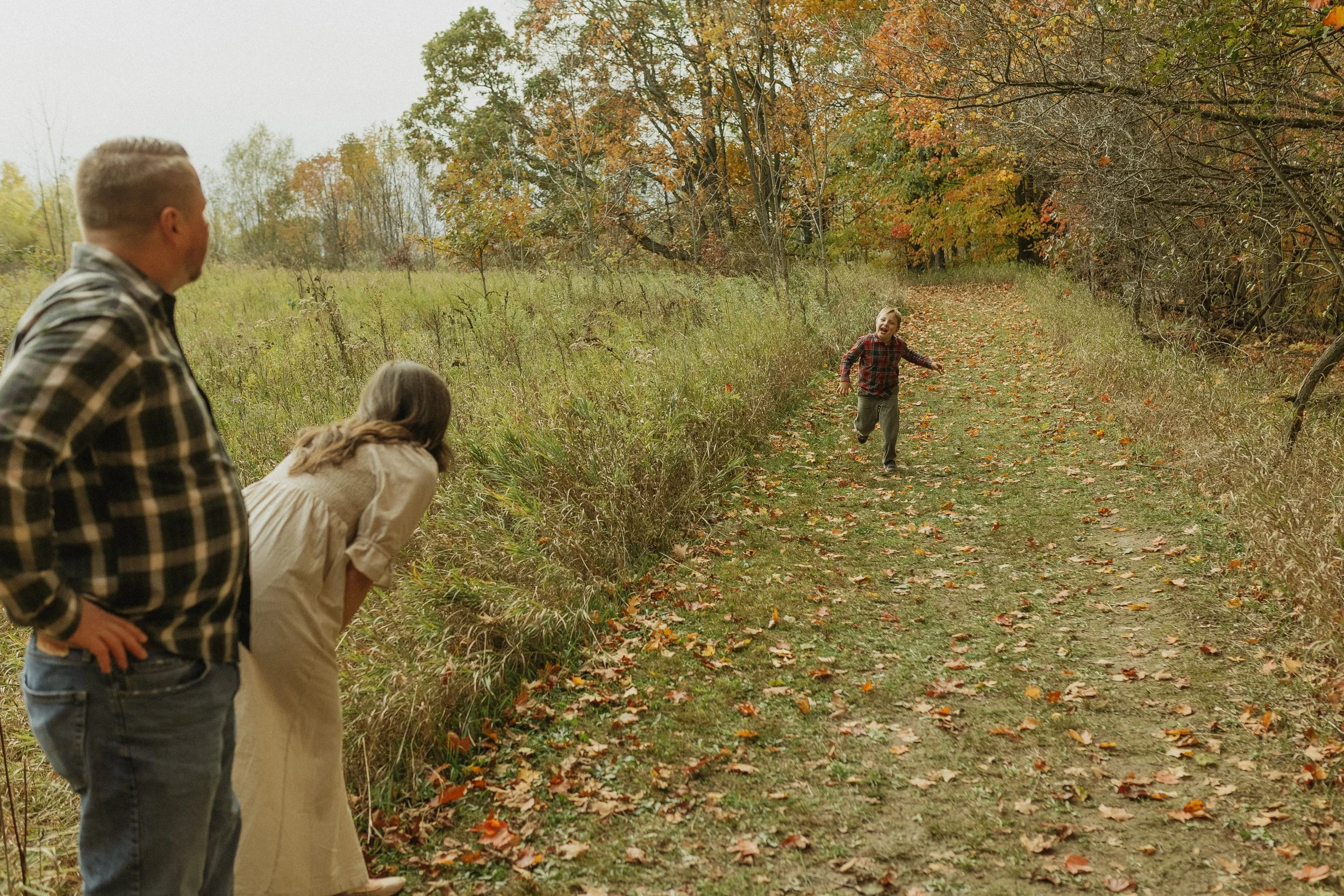 A young boy running towards two adults, a man and a woman, on a fall trail surrounded by trees with orange and yellow leaves.