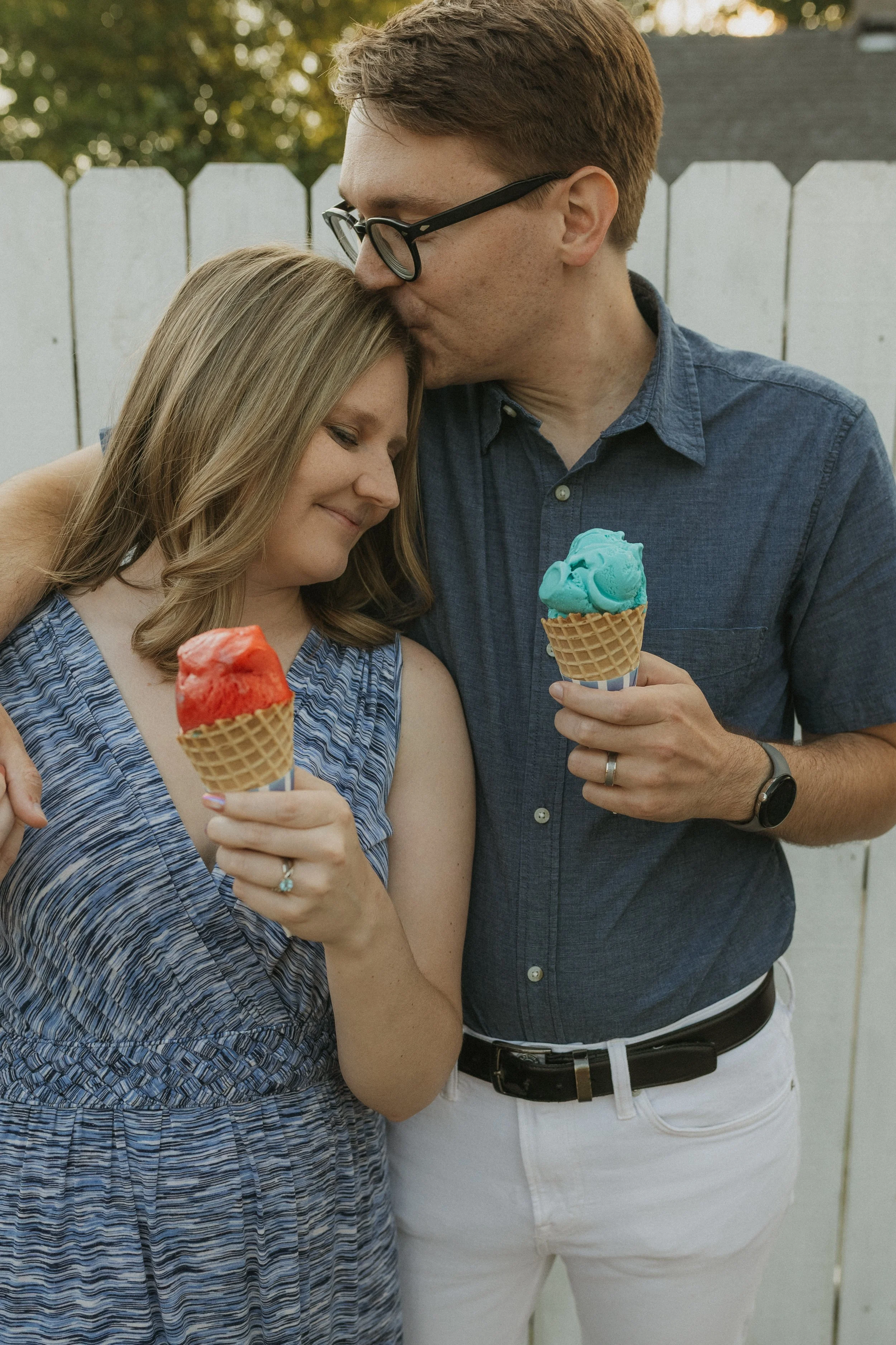A young couple holding ice cream cones, standing close together outdoors with a white fence in the background. The woman has light brown hair and is smiling with her eyes closed, holding a red ice cream cone. The man has glasses, short red hair, and 