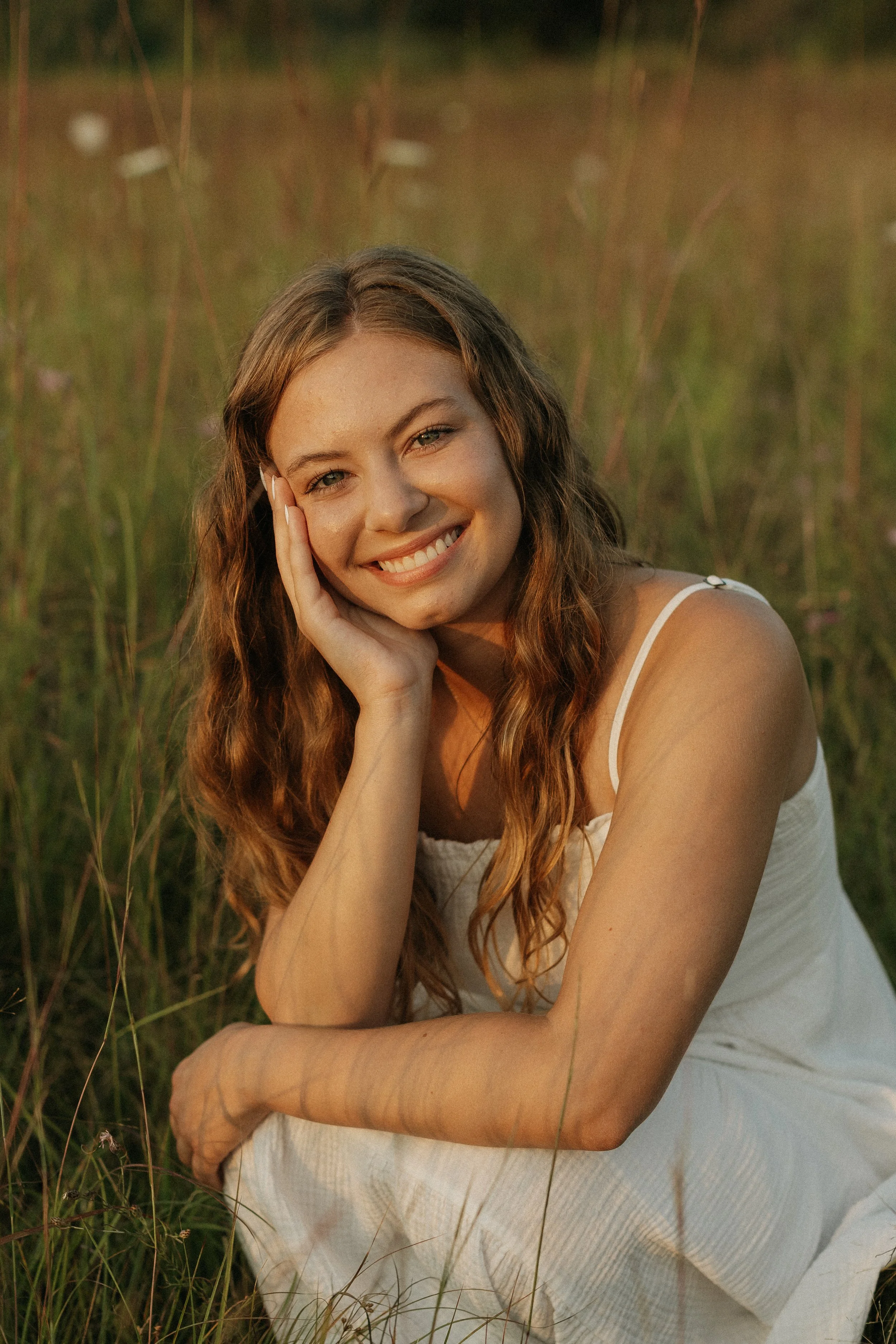 A young woman with long, wavy hair and wearing a white dress, smiling and resting her head on her hand while sitting in a grassy field during sunset.
