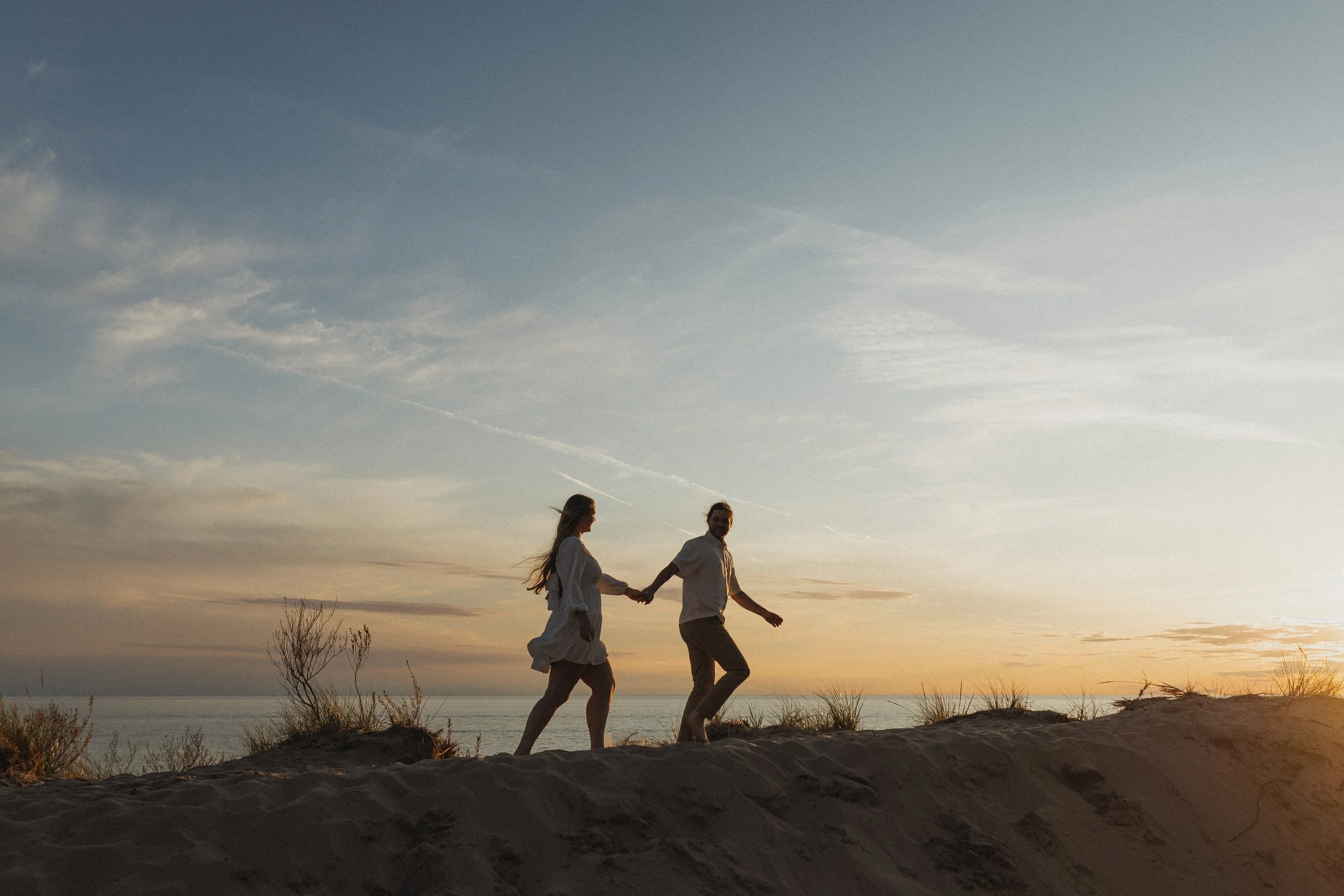 A couple walking hand in hand on a sandy beach at sunset, with the sky and ocean in the background.