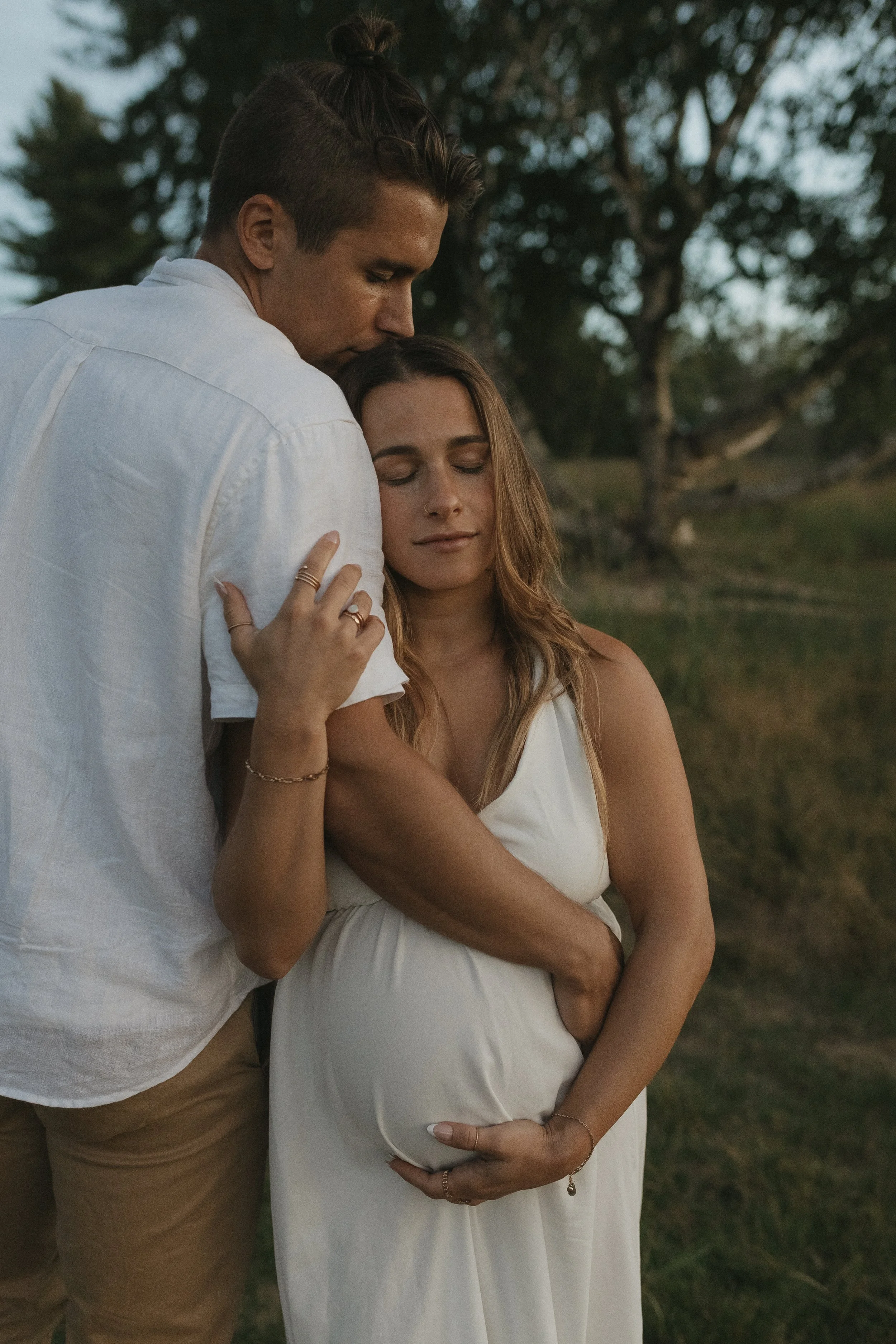 A man embracing a pregnant woman outdoors, both with serene expressions and eyes closed, during dusk or evening.
