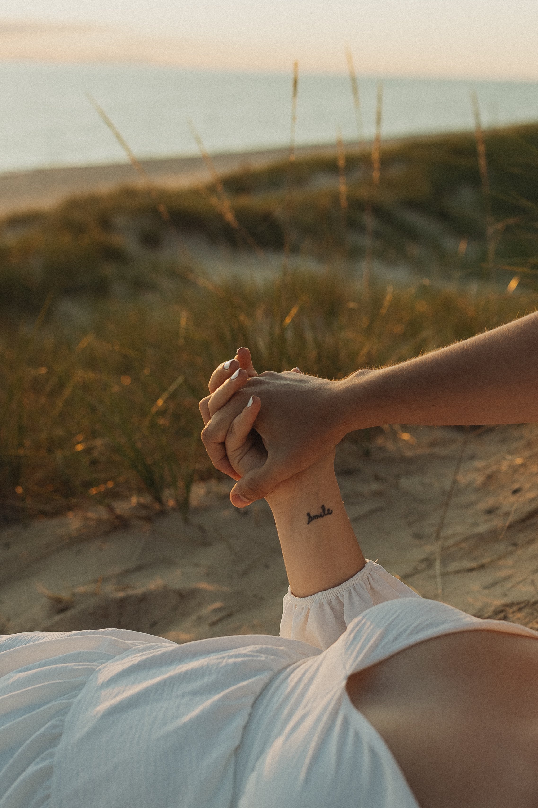 Hands intertwined on a sandy beach during sunset, one hand with a tattoo that says 'smile', and the other holding it gently.