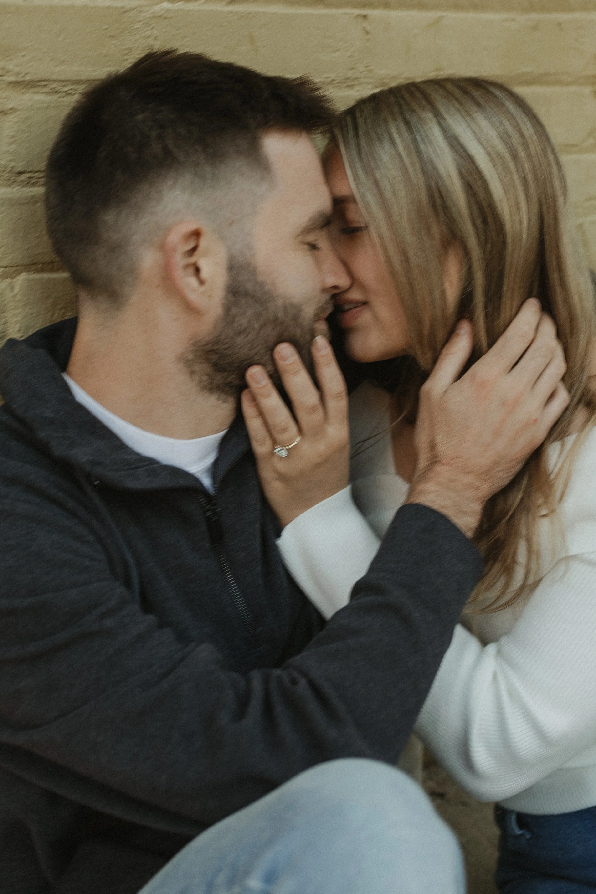 A close-up of a couple tenderly touching faces with their eyes closed, holding each other's faces gently in front of a brick wall.