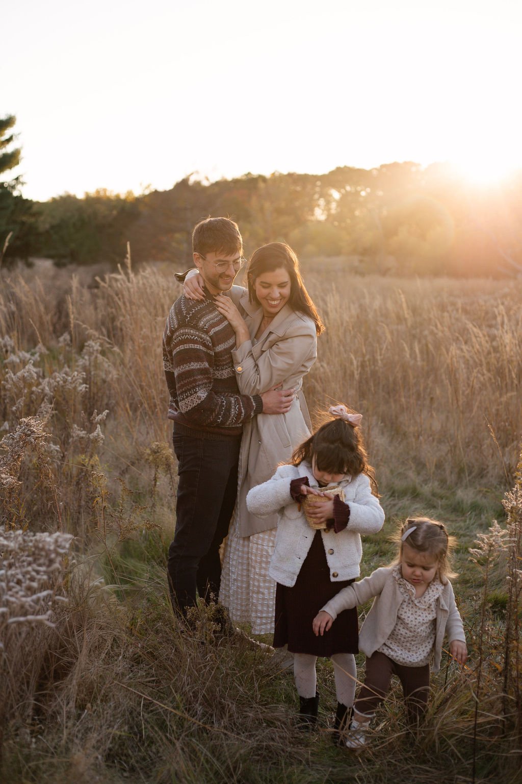 A happy family of four, including two young girls and their parents, embracing and walking through a field of tall grass at sunset.