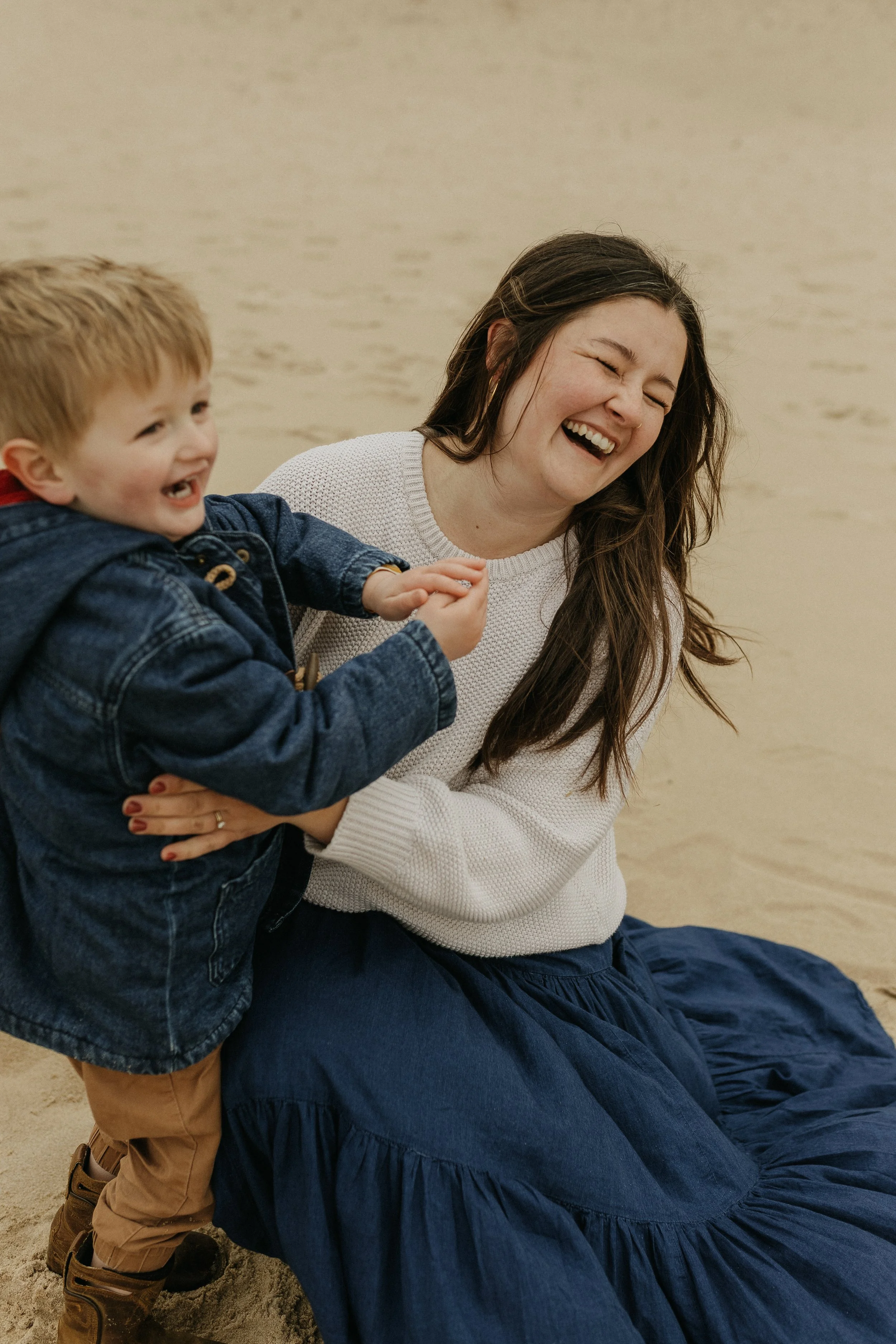 A woman and a young boy laughing and playing together on the sand at a beach.