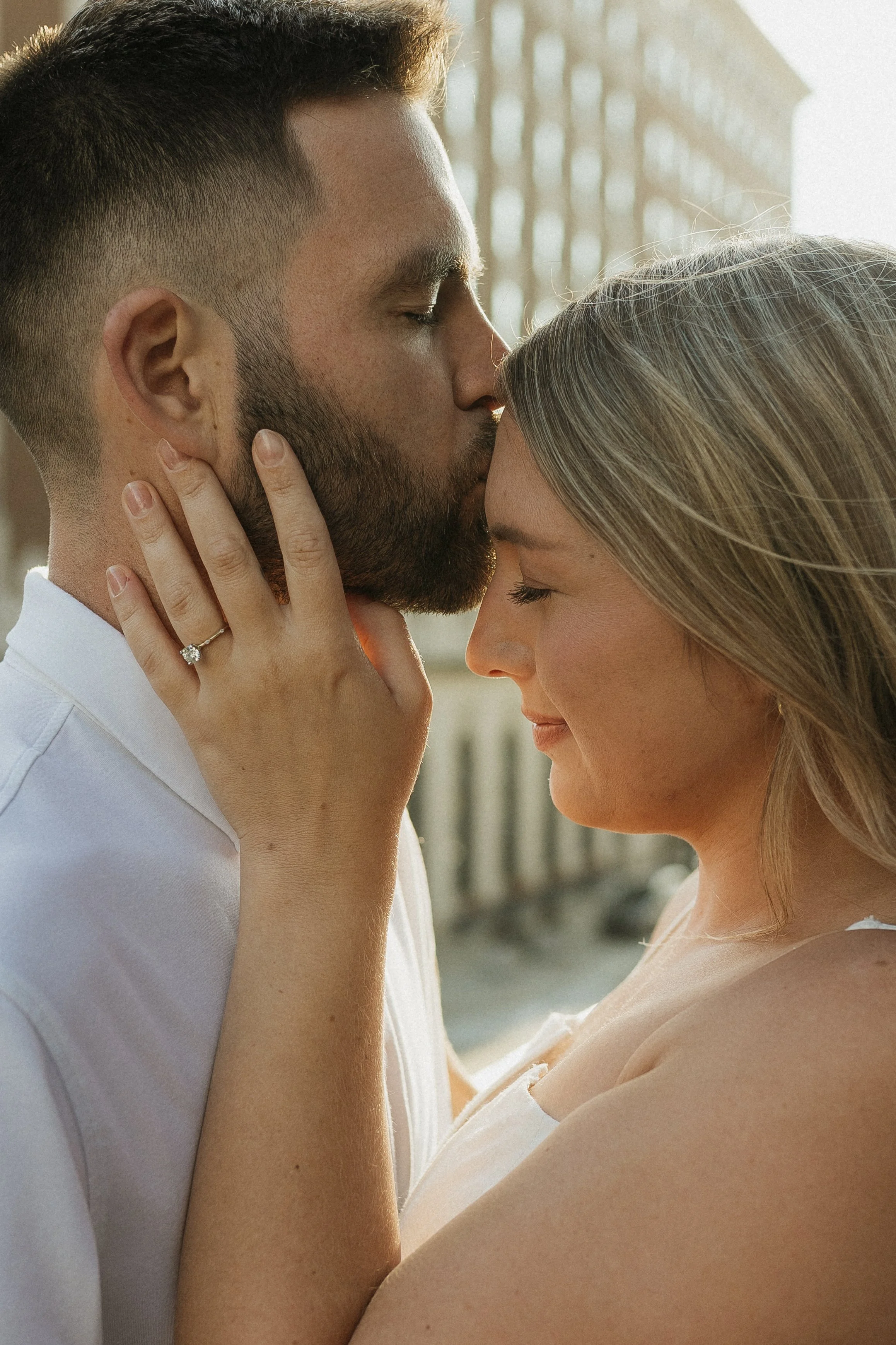A man and woman with closed eyes sharing a tender moment, with the man kissing the woman's forehead; she is wearing a ring on her left hand.