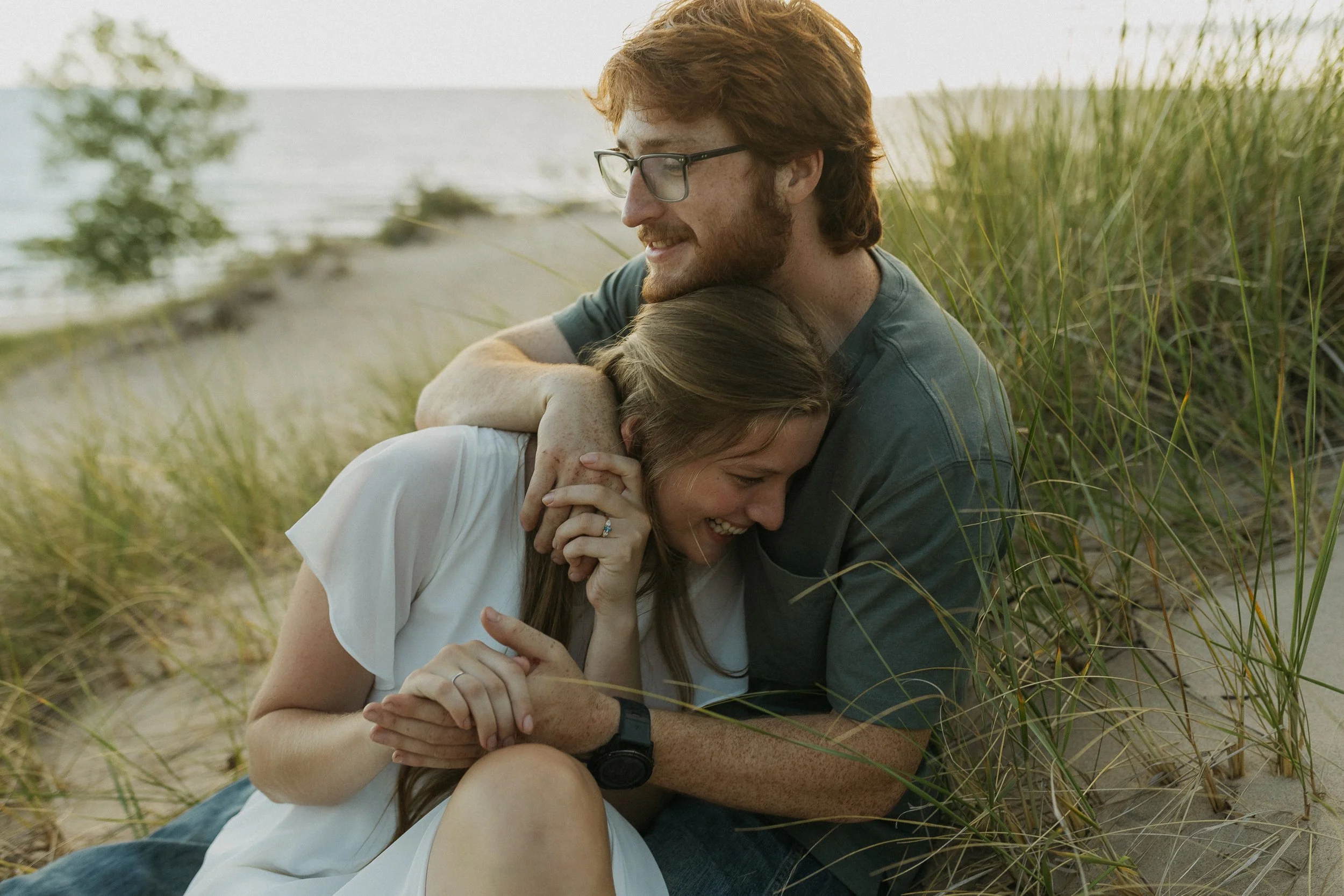 A young couple sitting on the beach, embracing and laughing together, surrounded by sand dunes and tall grass.