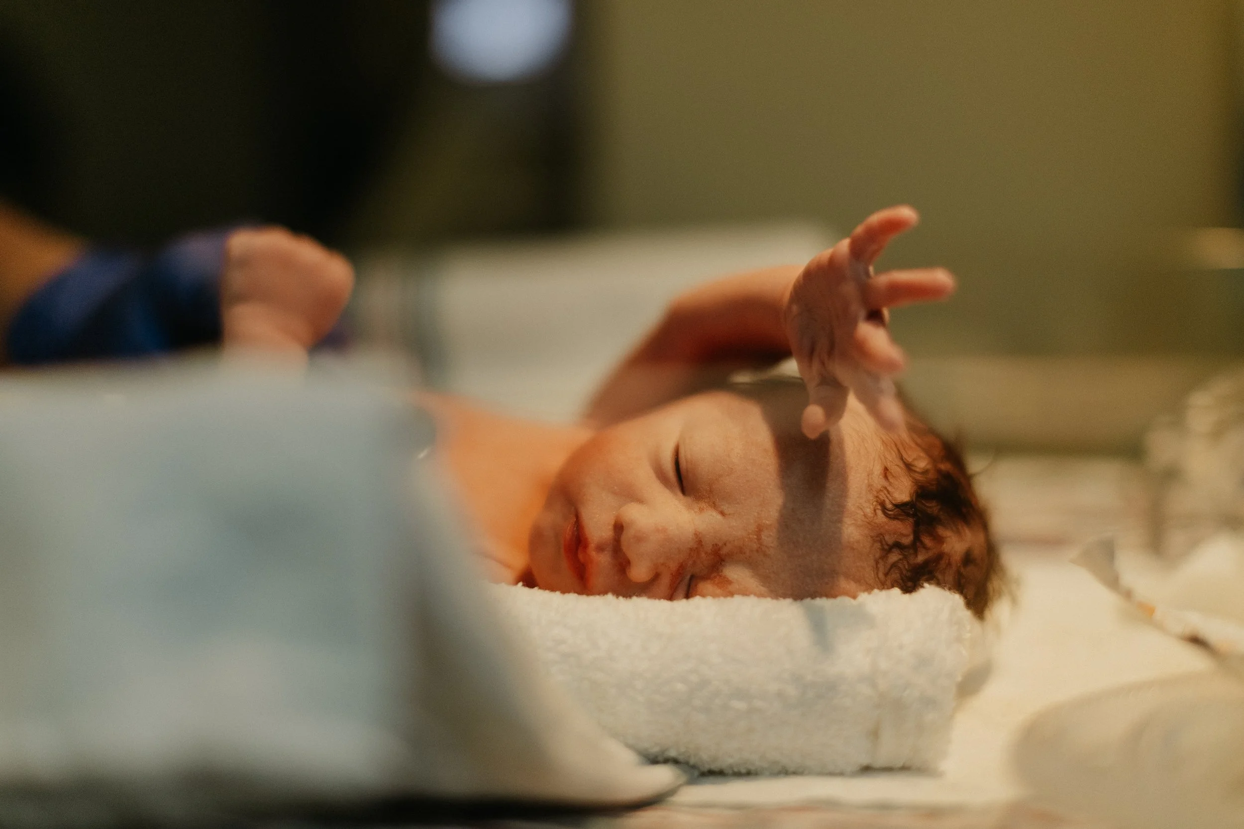 Close-up of a newborn baby lying on a white blanket, with eyes closed and a hand raised above the face.