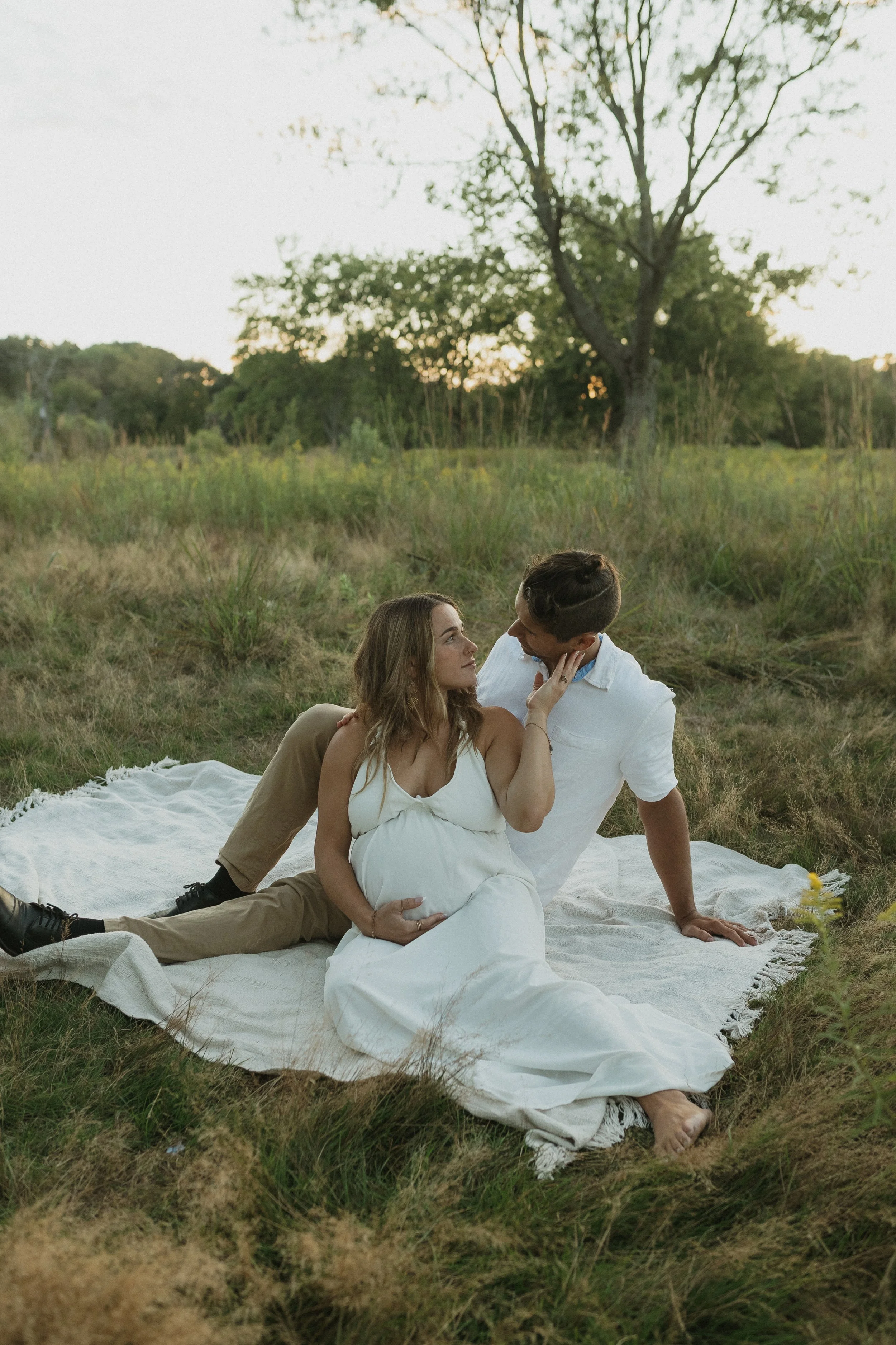 A pregnant woman and her partner sit on a white blanket in a grassy field, sharing an intimate moment at sunset.