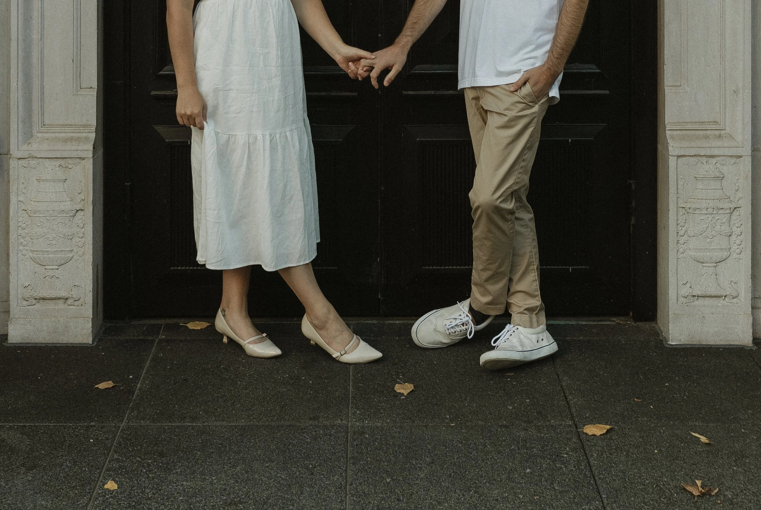 A couple holding hands in front of a black door, seen from the waist down, with the woman's white dress and beige heels and the man's beige pants and white sneakers visible.