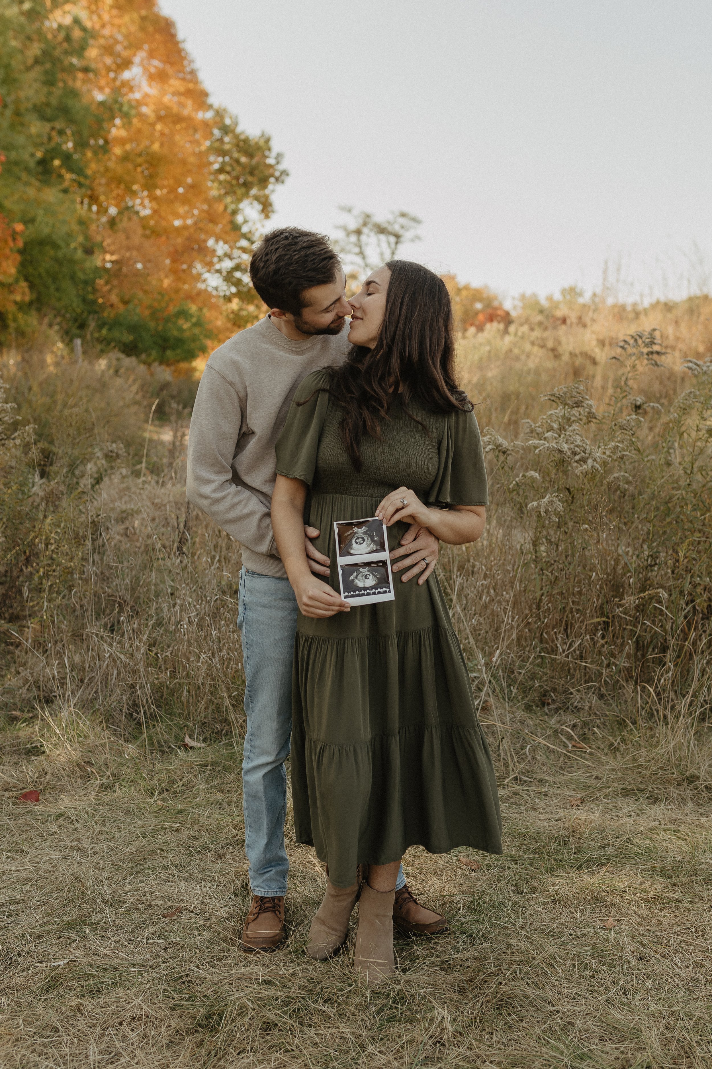 Couple posing outdoors holding ultrasound photos, surrounded by autumn foliage.