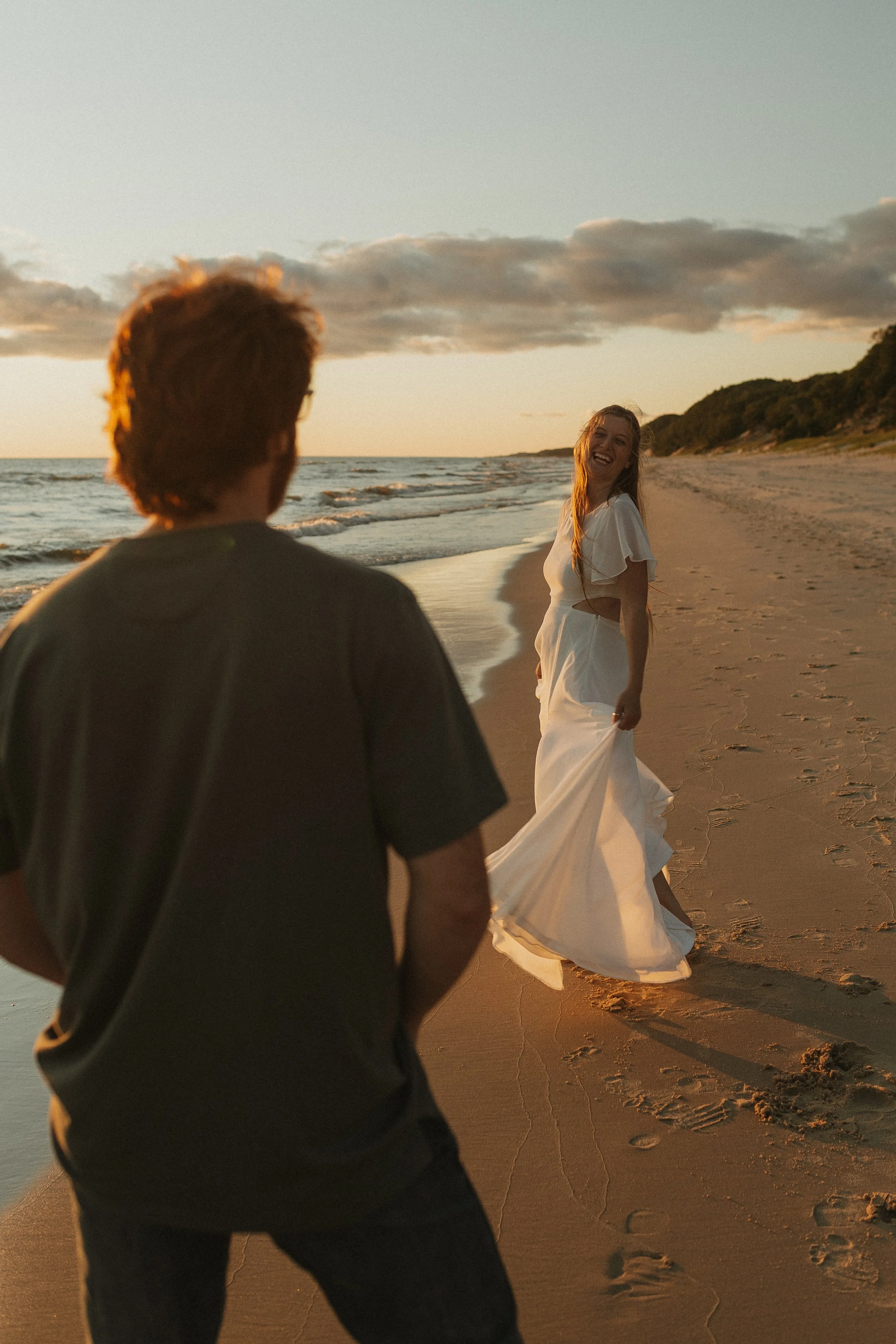 A woman in a white dress on a beach at sunset, smiling and looking at a man with his back to the camera.