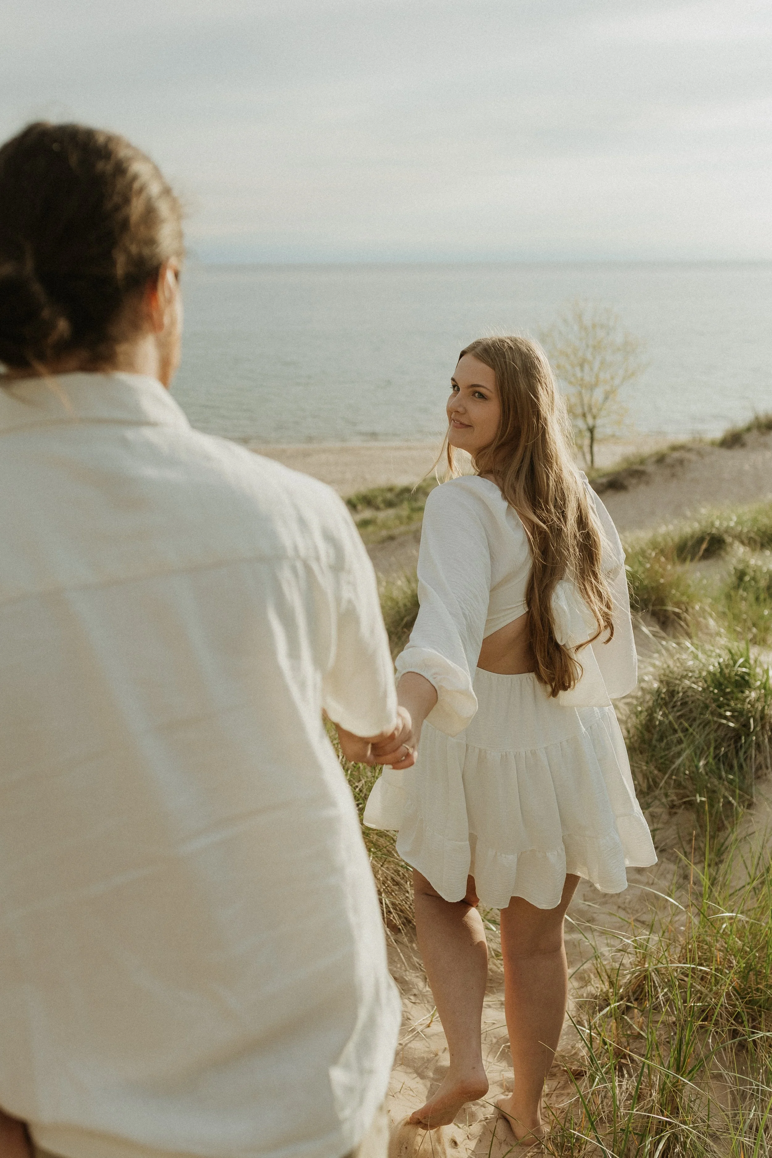 A young woman with long hair in a white dress holding hands with a man on a beach near the water, with grass and sand dunes around them.