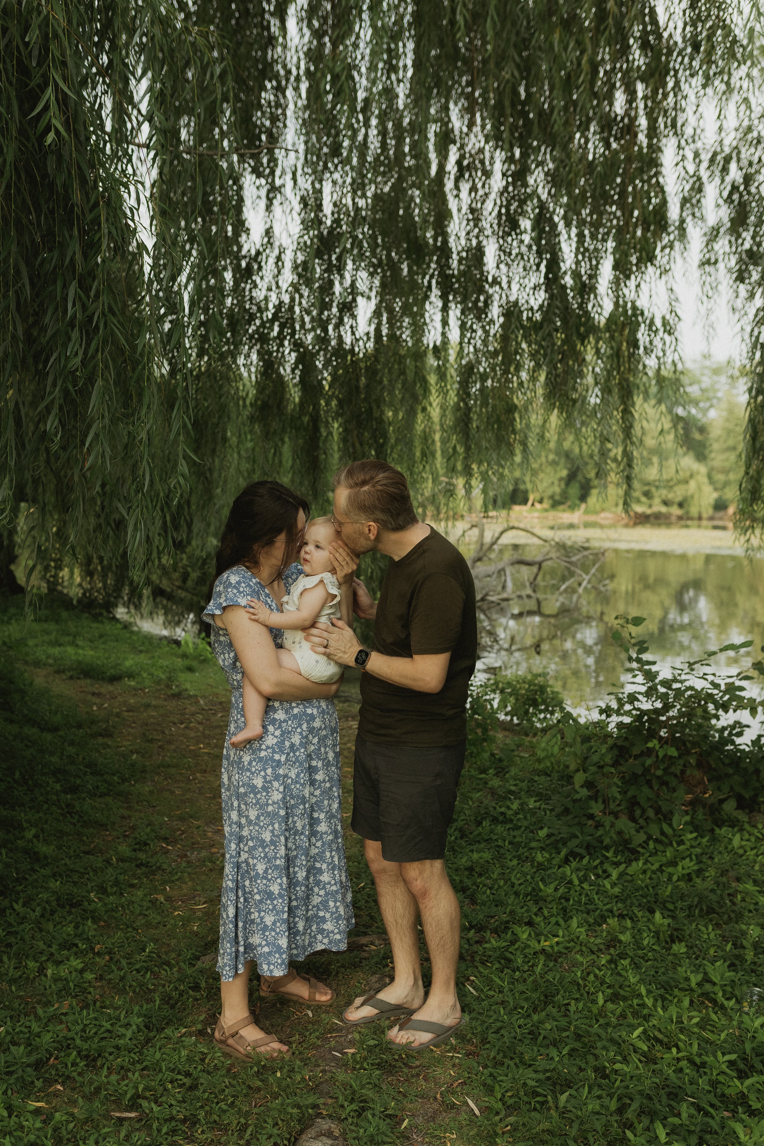Family of three enjoying time by a lake under a large weeping willow tree, with the father kissing the child's forehead, all smiling.