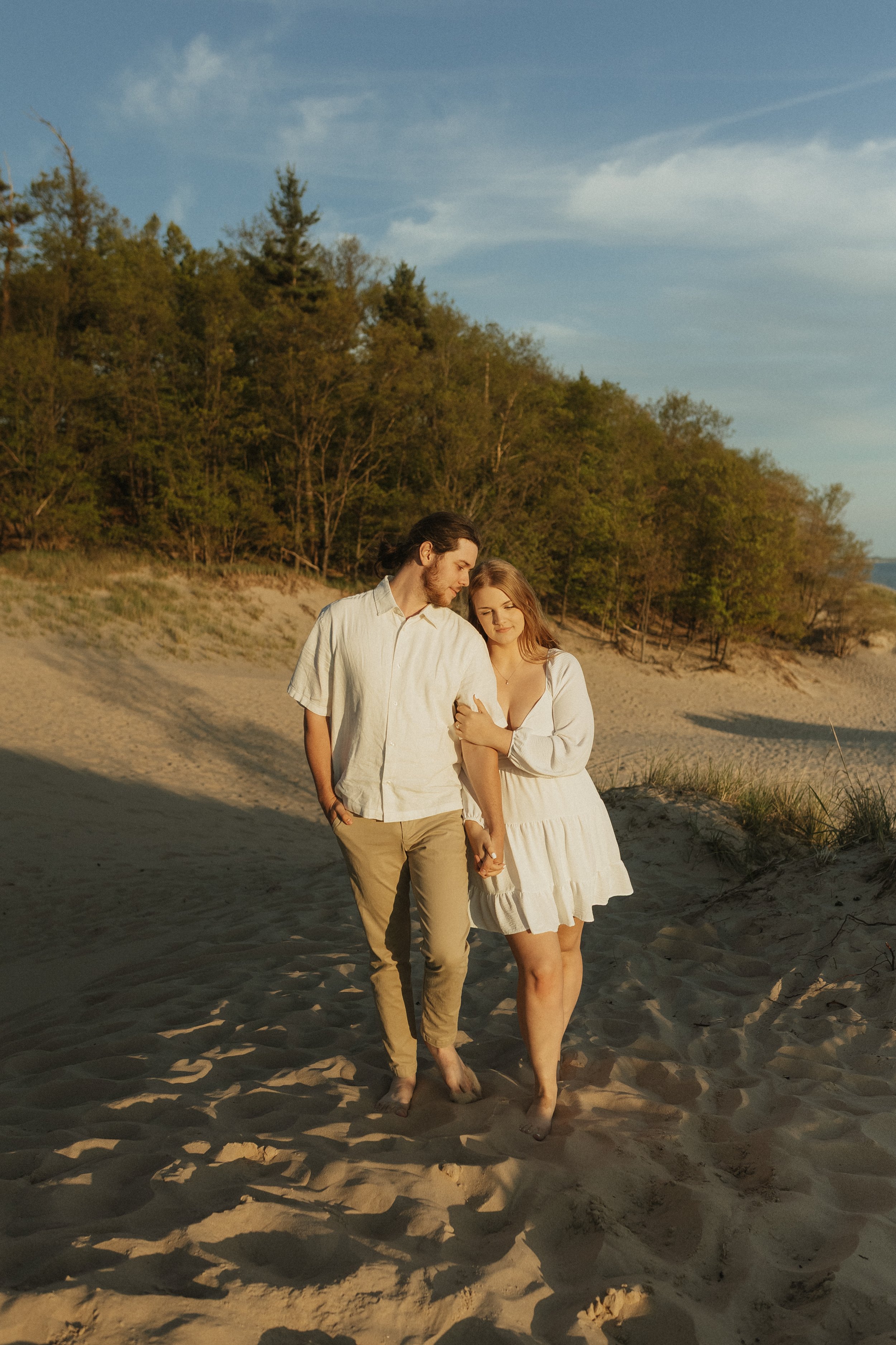A couple walking on a sandy beach holding hands, with trees and a blue sky in the background during sunset.