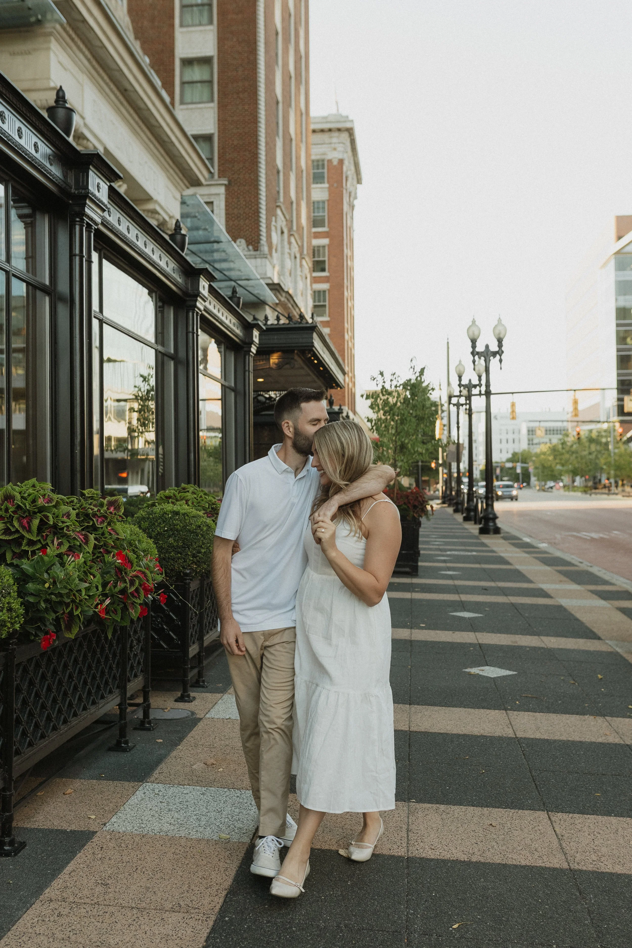 A couple walking and hugging on a city sidewalk in front of a restaurant or cafe with large windows and outdoor plants, with buildings and street lamps in the background.