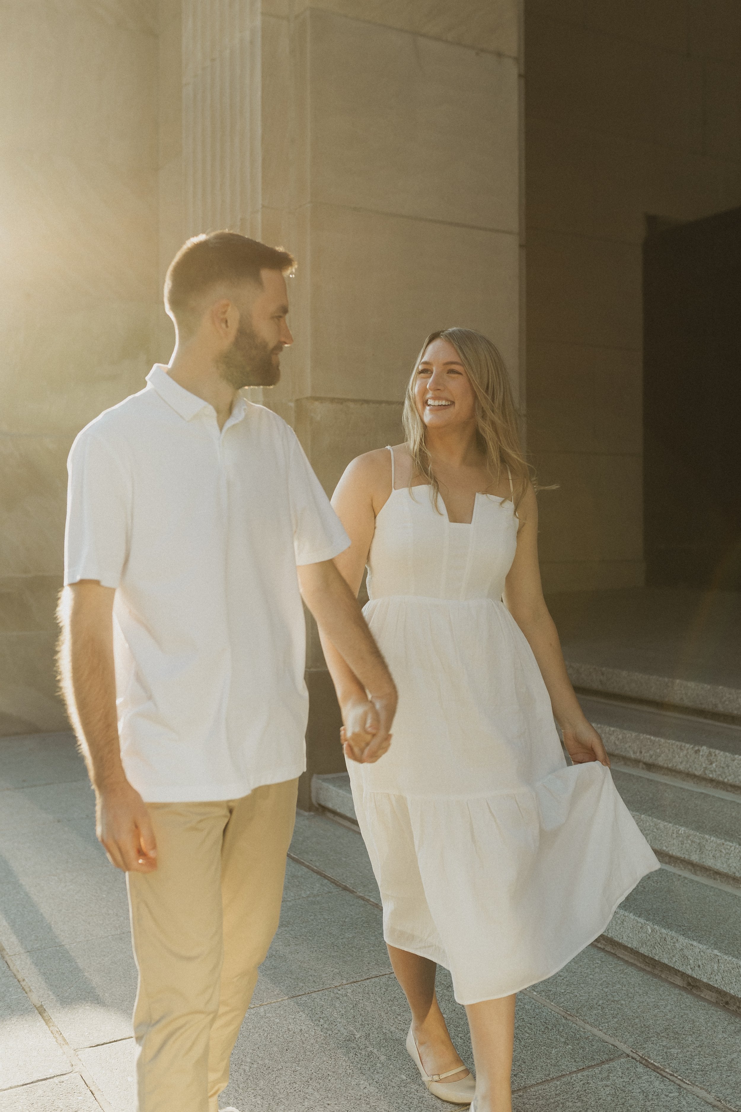 A young couple holding hands and walking outdoors, bathed in warm sunlight. The woman is wearing a white dress, smiling and looking at the man. The man is wearing a white shirt and beige pants, looking at the woman.