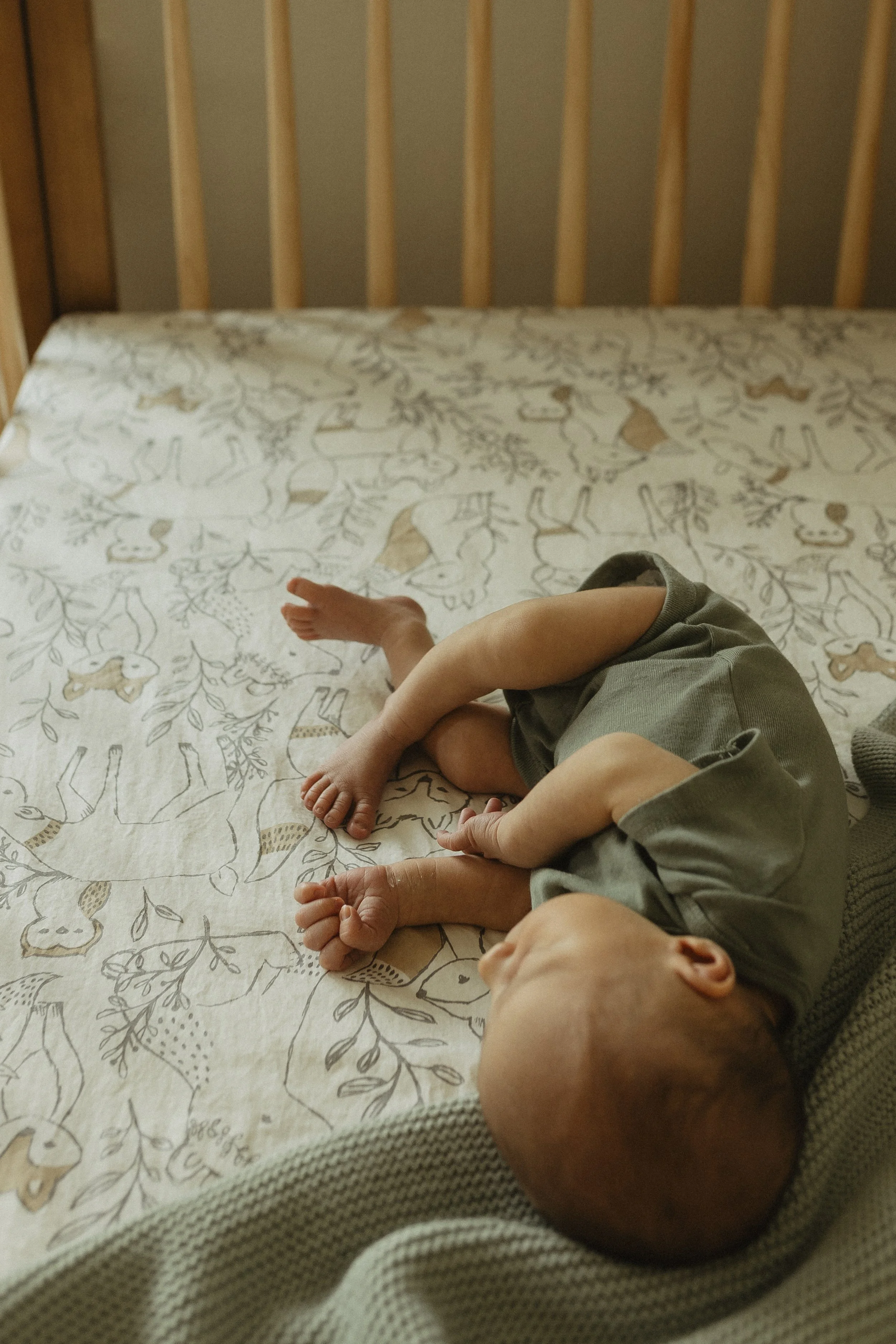 A baby lying on a bed with a deer and woodland creature pattern, curled up on their side with arms and legs bent.
