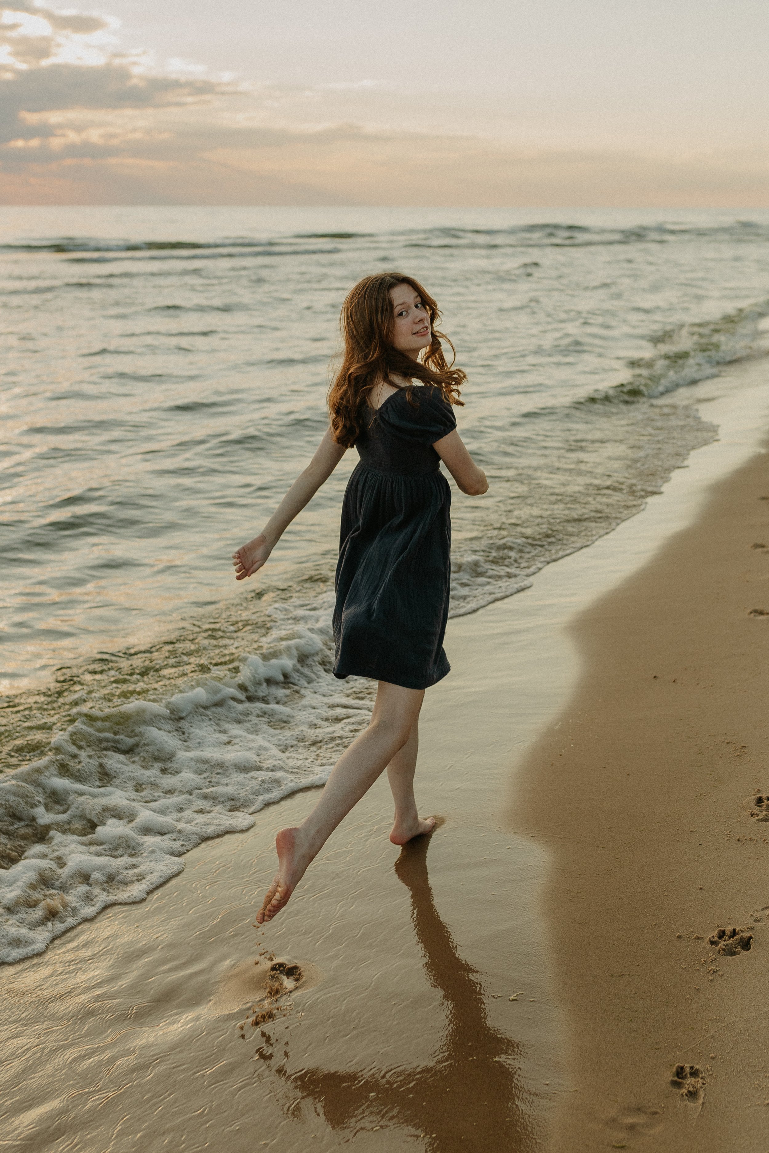 A woman in a black dress walking barefoot along the shoreline at the beach during sunset.