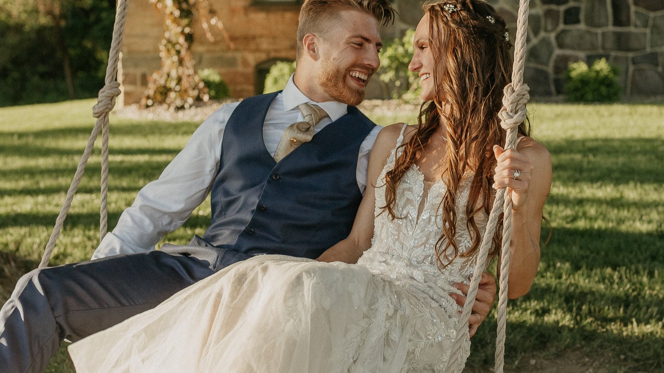 Smiling couple on a swing, dressed in wedding attire, outdoors with greenery and a stone building in the background.