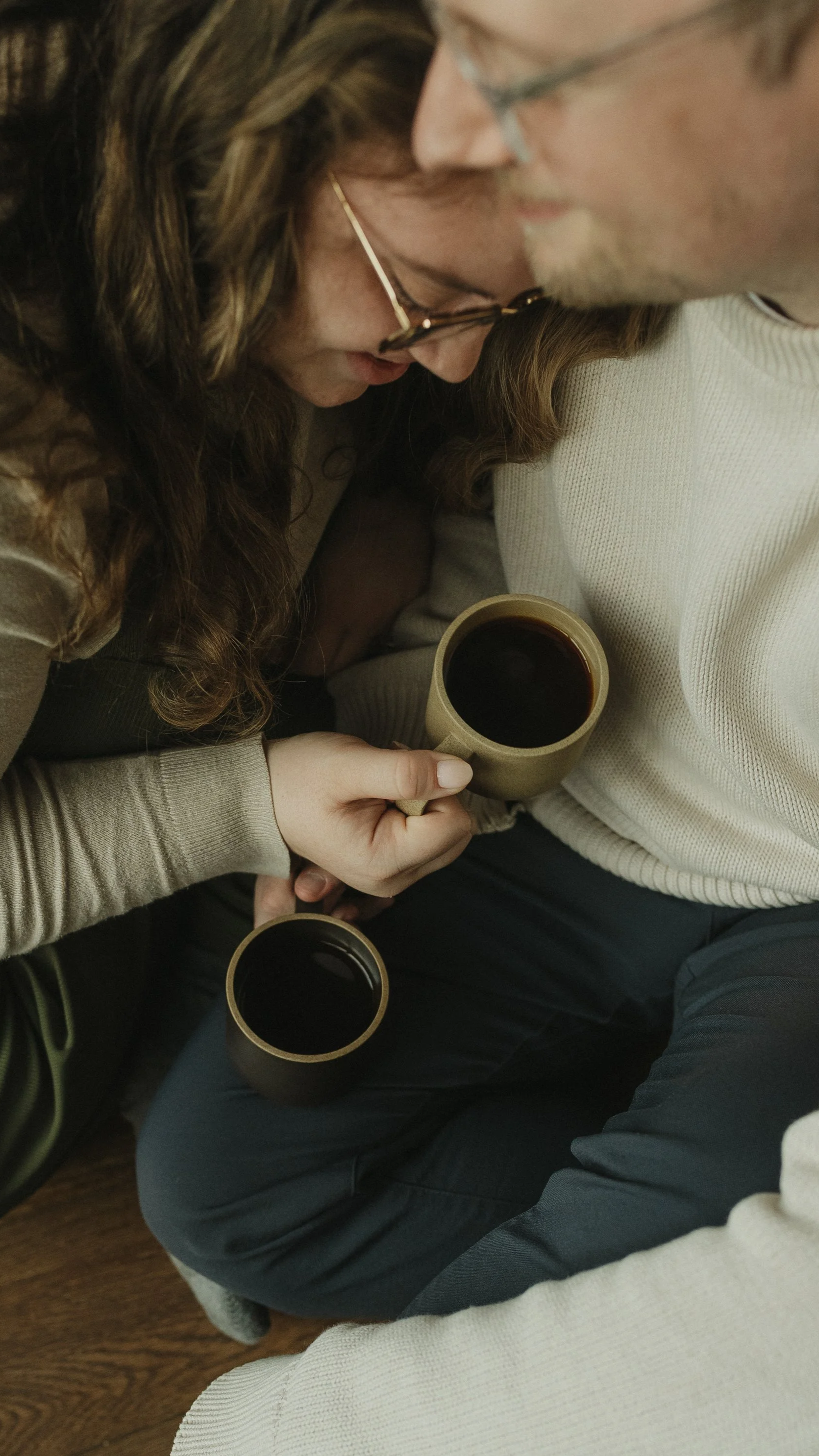 A woman and a man sitting close together on the floor at home, sharing a moment, each holding coffee mugs.