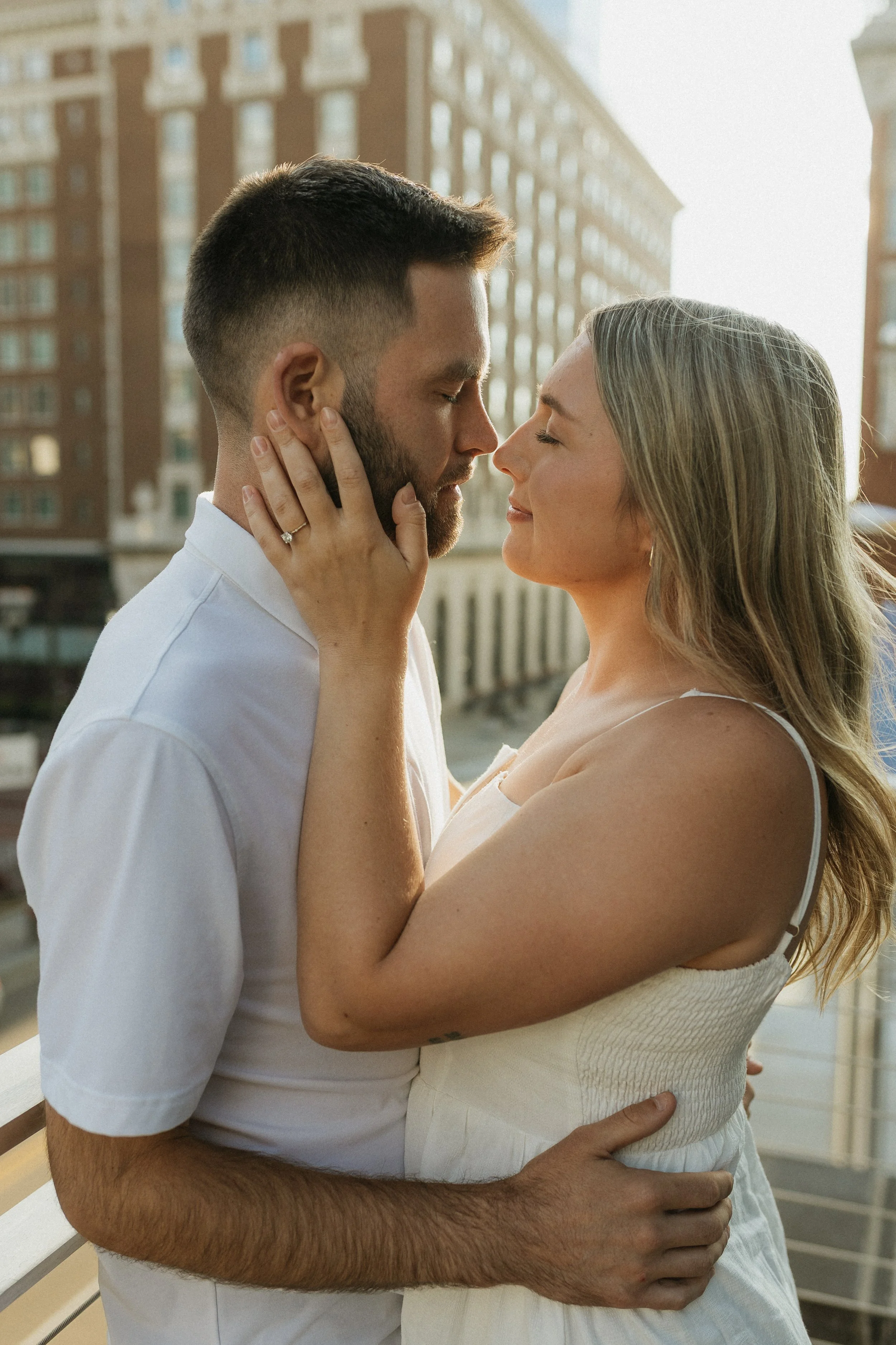 A couple standing close together on a balcony, with their faces almost touching and eyes closed, in an urban setting with buildings in the background.