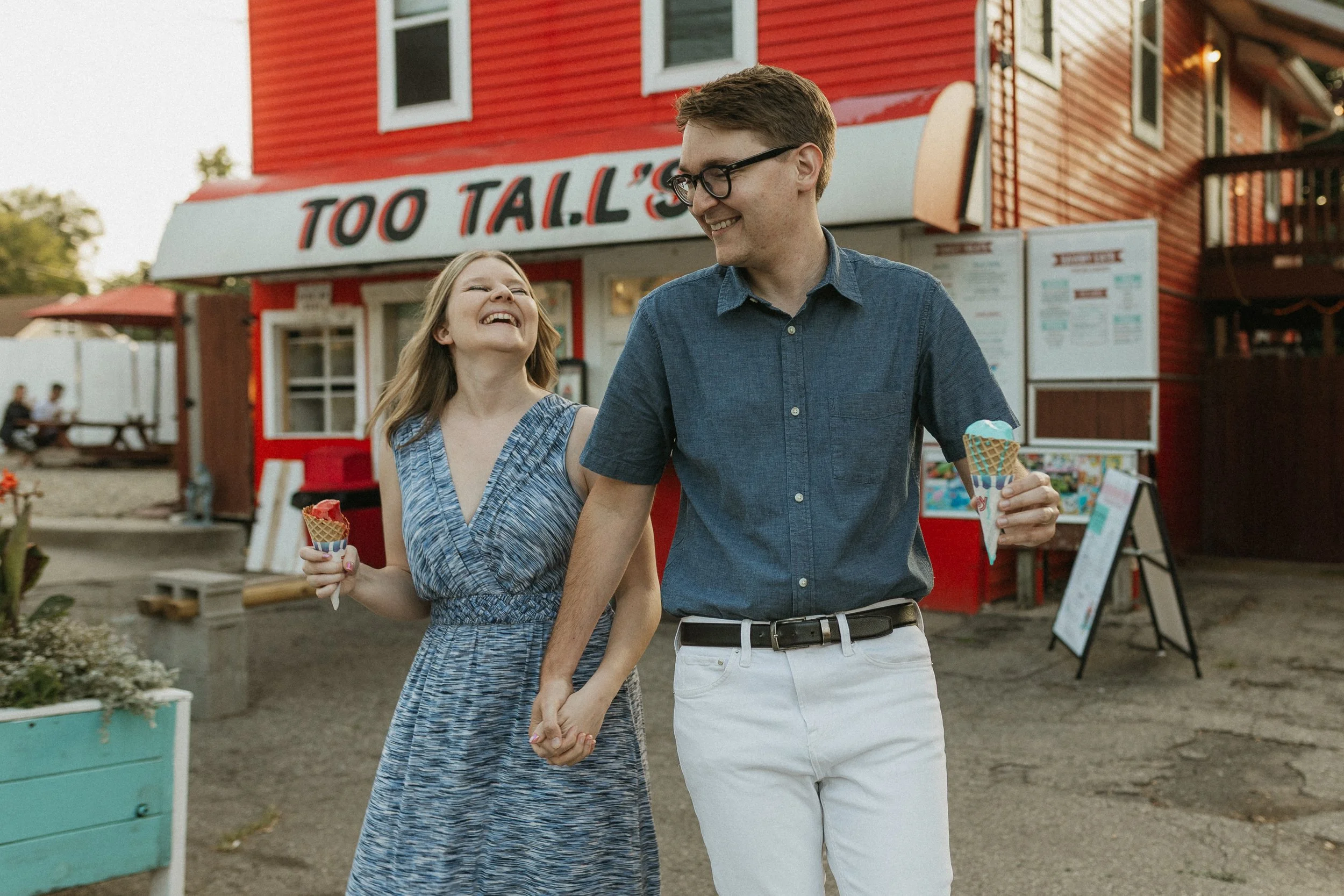 A smiling young woman and man holding hands and ice cream cones walking outside near a red ice cream stand that reads 'Too Tall's'.