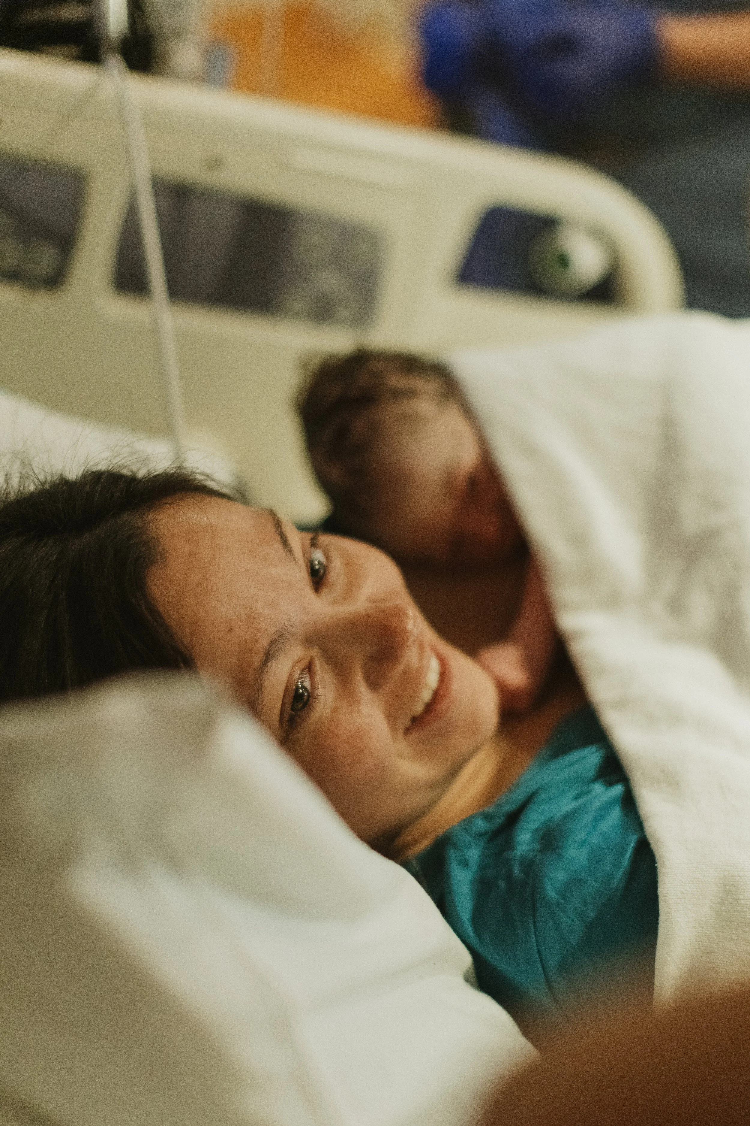 A woman lying in a hospital bed with a smiling face, holding her newborn baby partially covered with a white blanket.