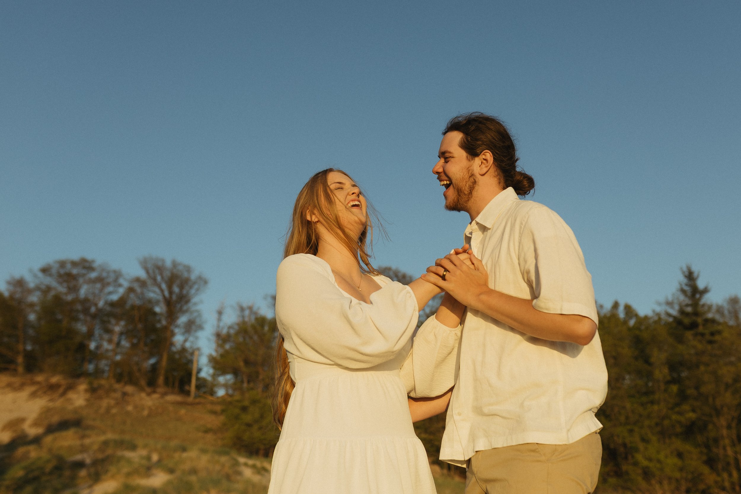 A joyful couple dancing outdoors during sunset, smiling and holding hands, with trees and a clear blue sky in the background.