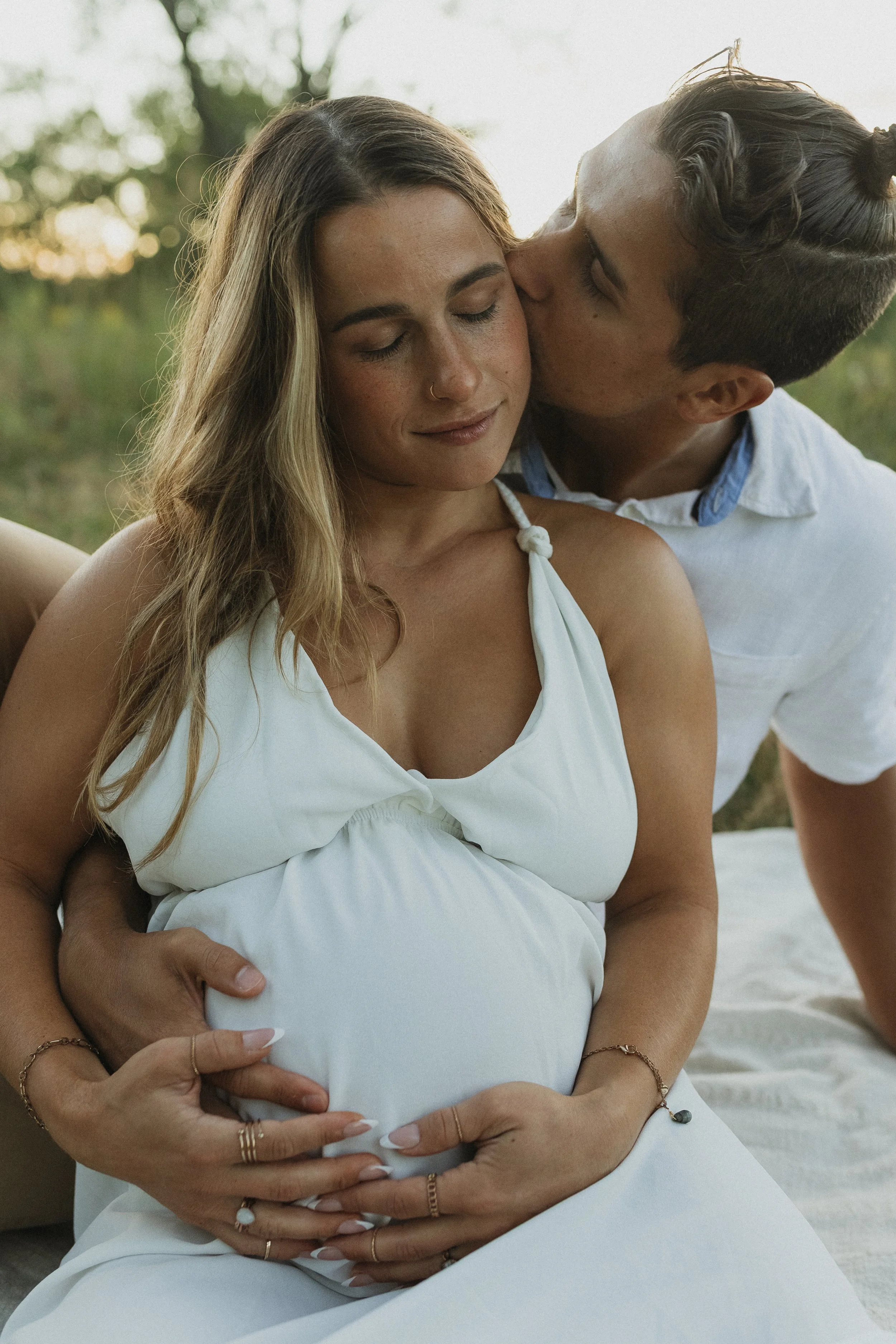 A pregnant woman in a white dress with jewelry, sitting outdoors with her partner, who is kissing her cheek. The woman is gently holding her pregnant belly with her hands.